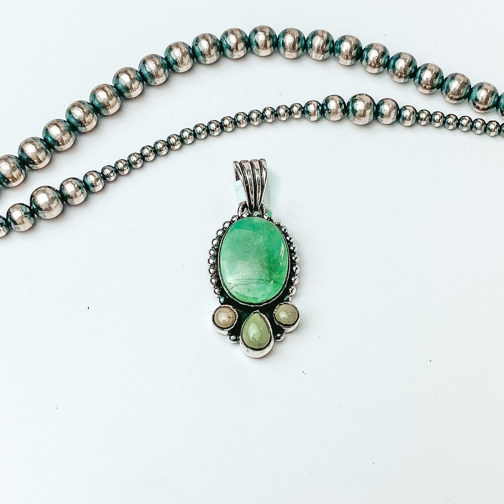 Centered in the picture is a turquoise round pendant with three accent stones on the bottom. Navajo pearls are laid above the pendant, all on a white background. 