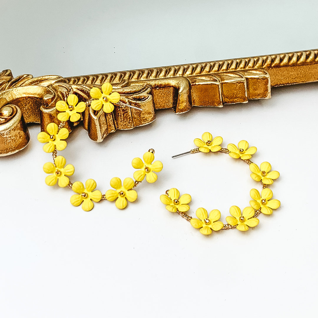 Gold hoop earrings with light yellow flower charms outlining the earrings. These flowers have a gold bead center. One hoop is pictured laying against a gold mirror while the other hoop is laying flat on the white background. 