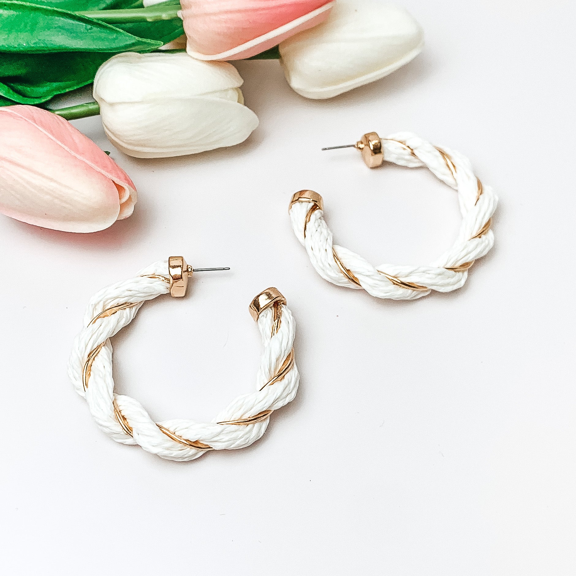 Pictured are white raffia twisted hoop earrings with gold detailing.  They are pictured with pink and white tulips on a white background.