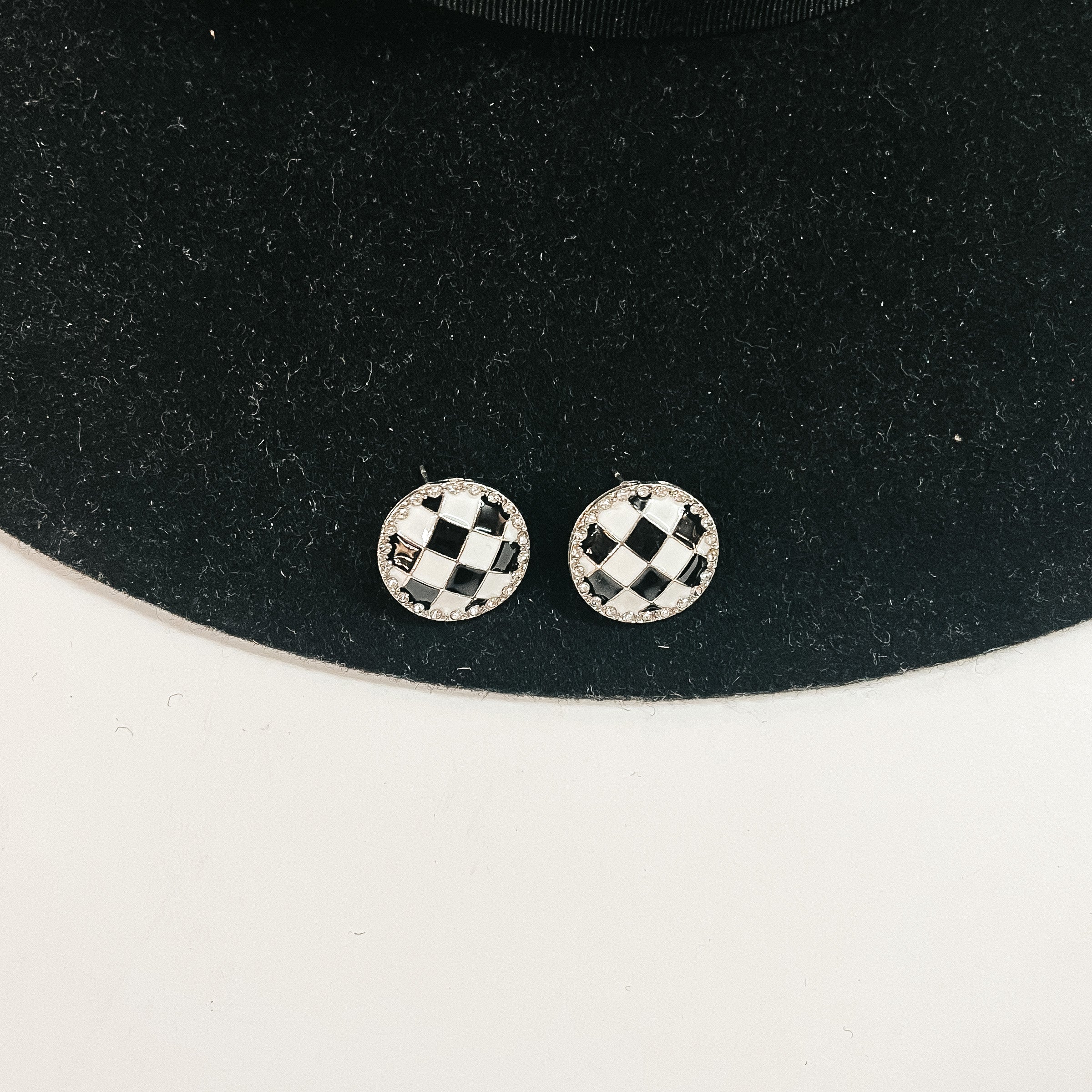 This is a pair of checkered patterned stud earrings in white/black in a gold  setting with clear crystals all around. This pair of earrings is laying on a  black felt hat brim and on a white background.