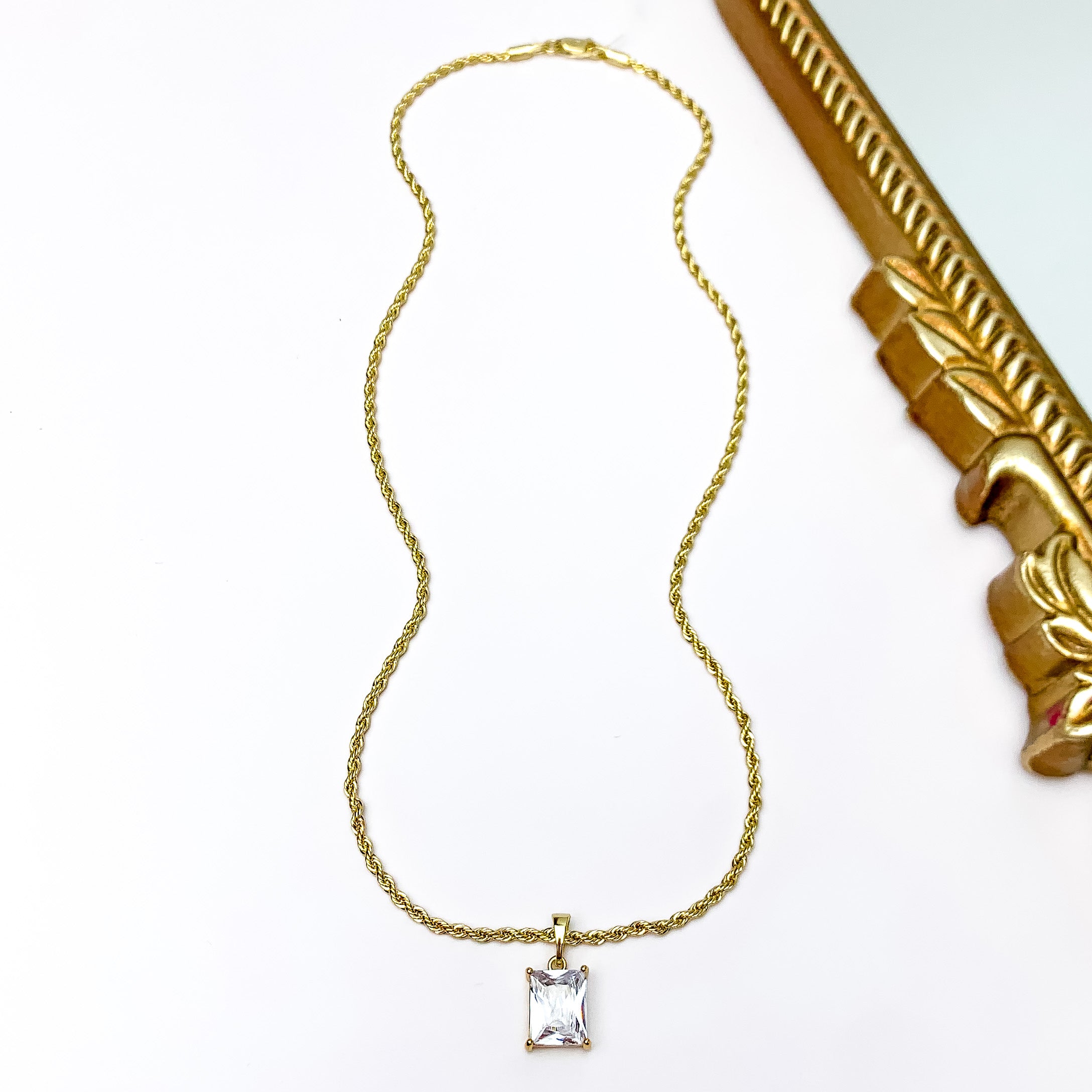 Pictured is a gold rope chain neckalce with a rectabgle, clear crystal pendant. This necklace is pictured on a white background with a gold mirror in the top right corner.  