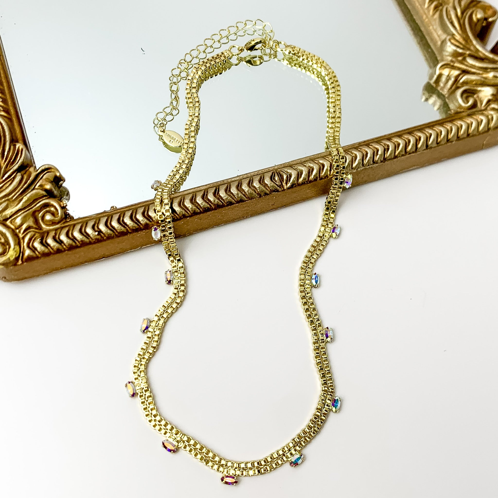 Pictured is a double, gold box chain necklace with ab crystal charms. This necklace is pictured partially laying on a gold mirror on a white background.    