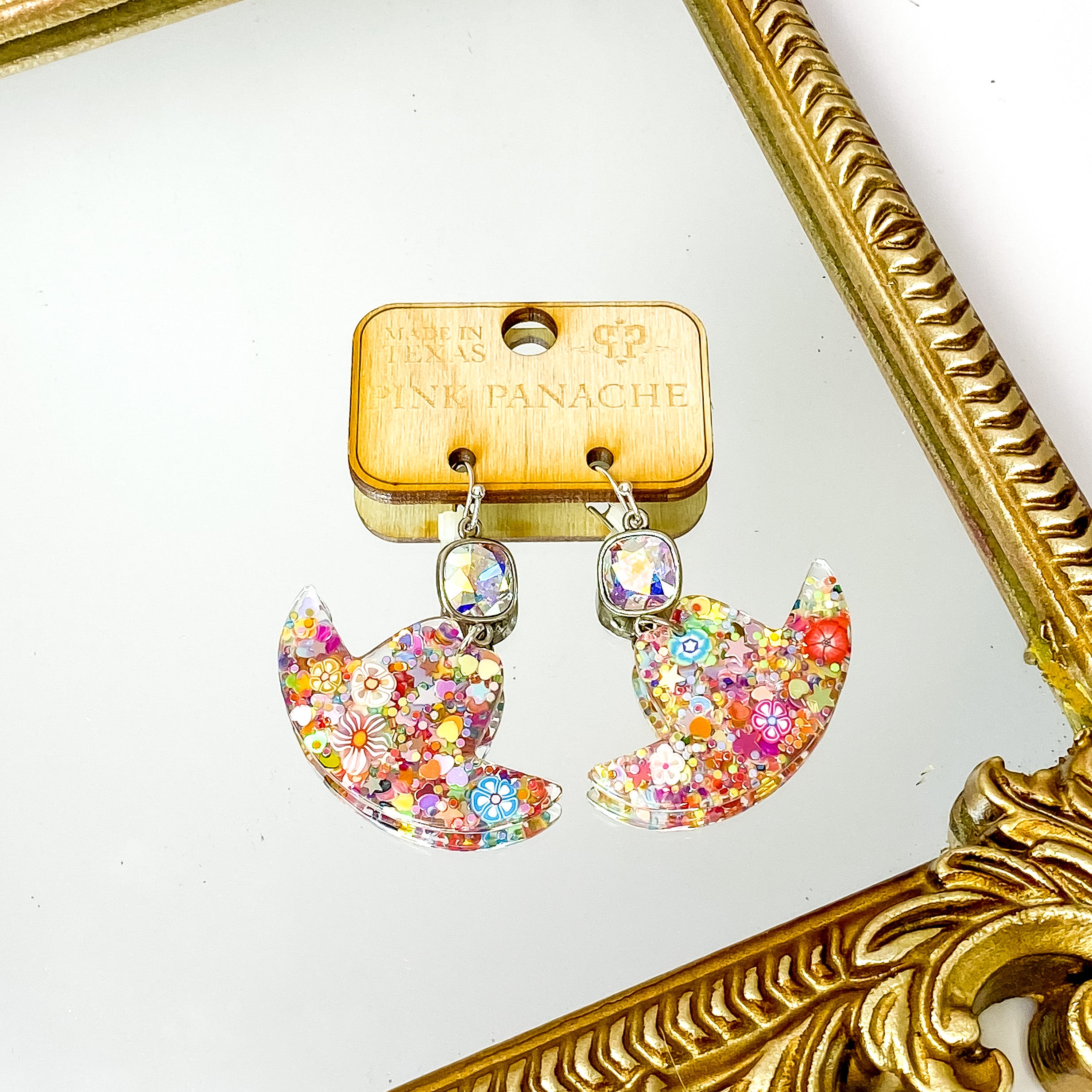 Silver, cushion cut AB crystal drop earrings with a hanging hat pendant. This pendant has multicolor glitter all over. These earrings are pictured on a wood earrings holder on a gold mirror on a white background. 
