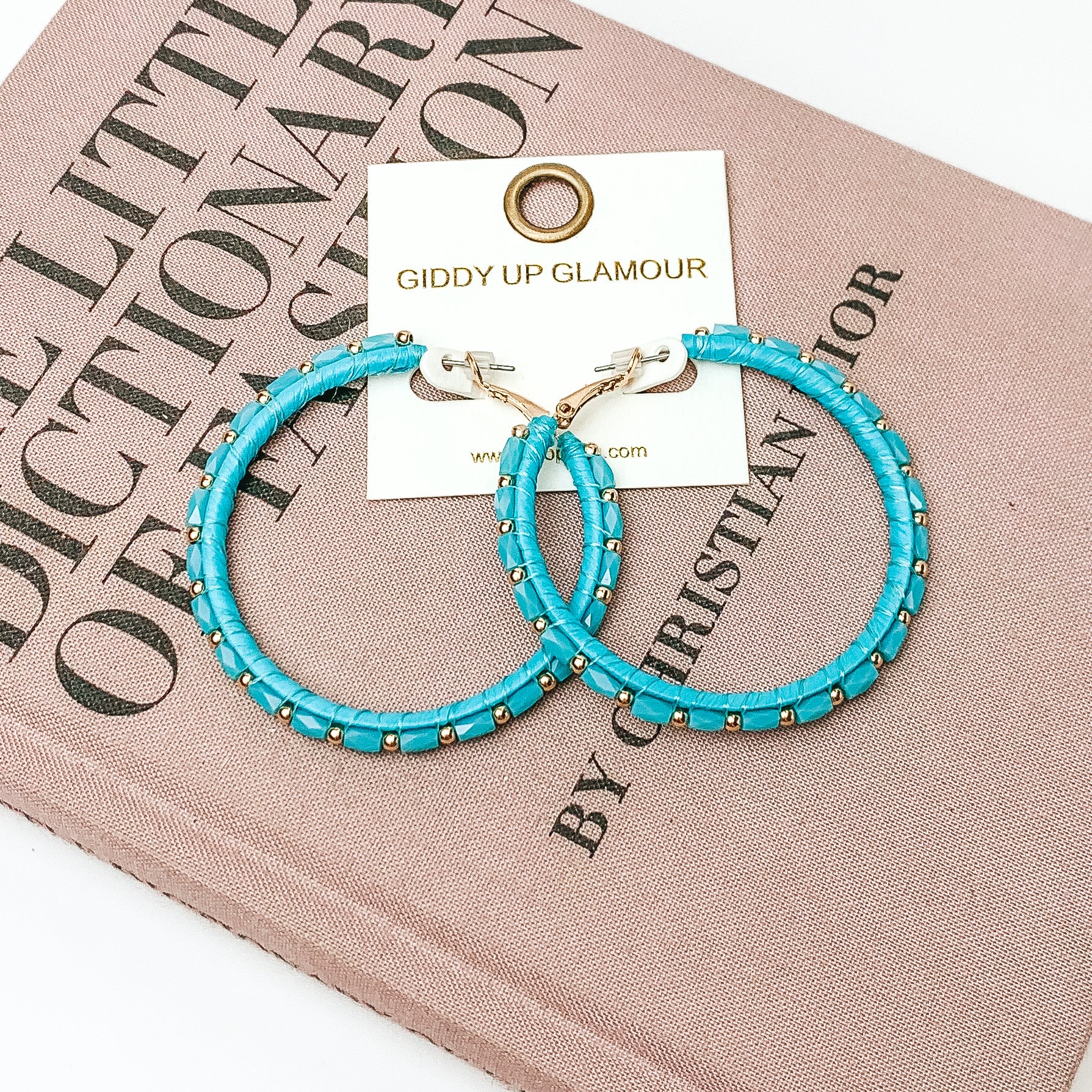 Pictured are circle beaded hoop earrings with gold spacers in turquoise. They are pictured with a pink fashion journal on a white background