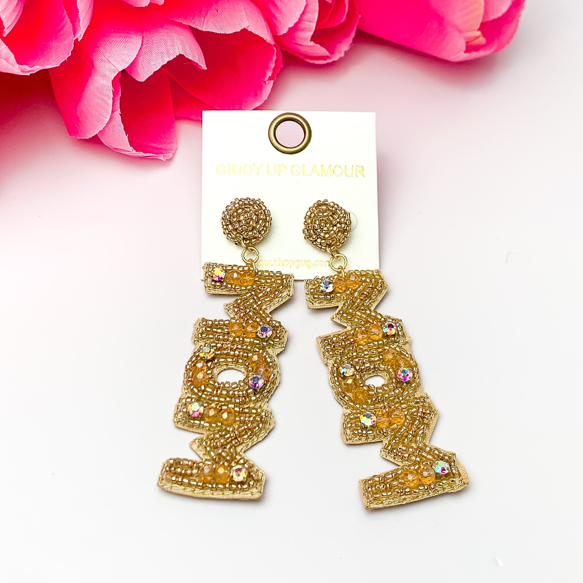 These are gold beaded and ab crystal earrings with a drop that says, MOM. These earrings are taken on a white background with pink flowers.