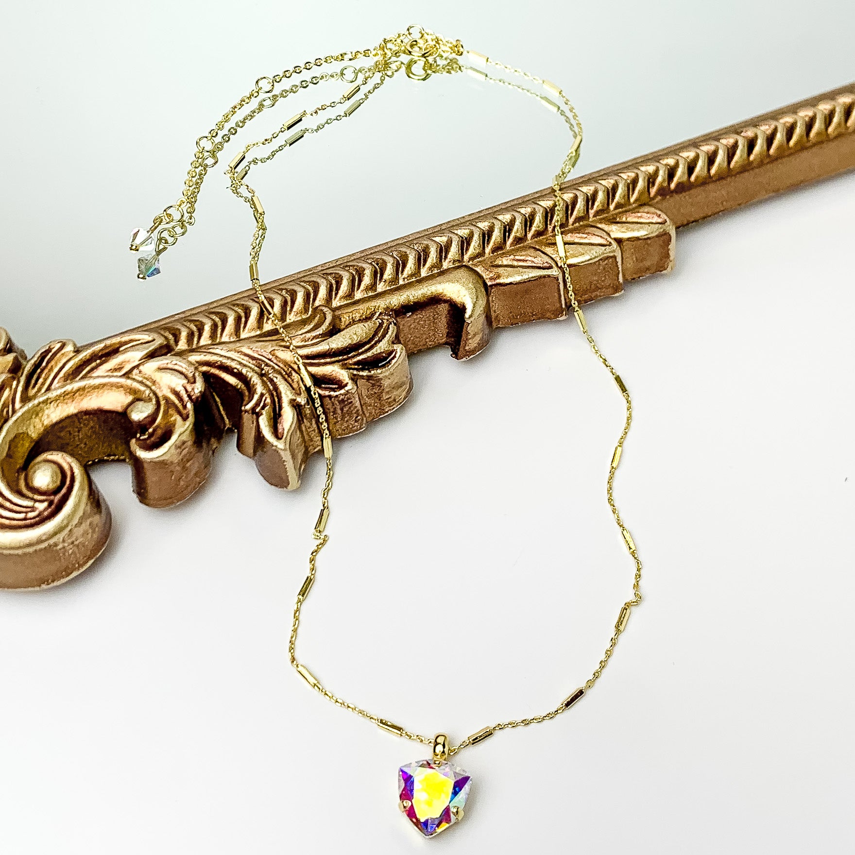 Pictured is a gold, bar chain necklace with a heart ab crystal pendant. This necklace is pictured partially Laying on a gold mirror on a white background. 