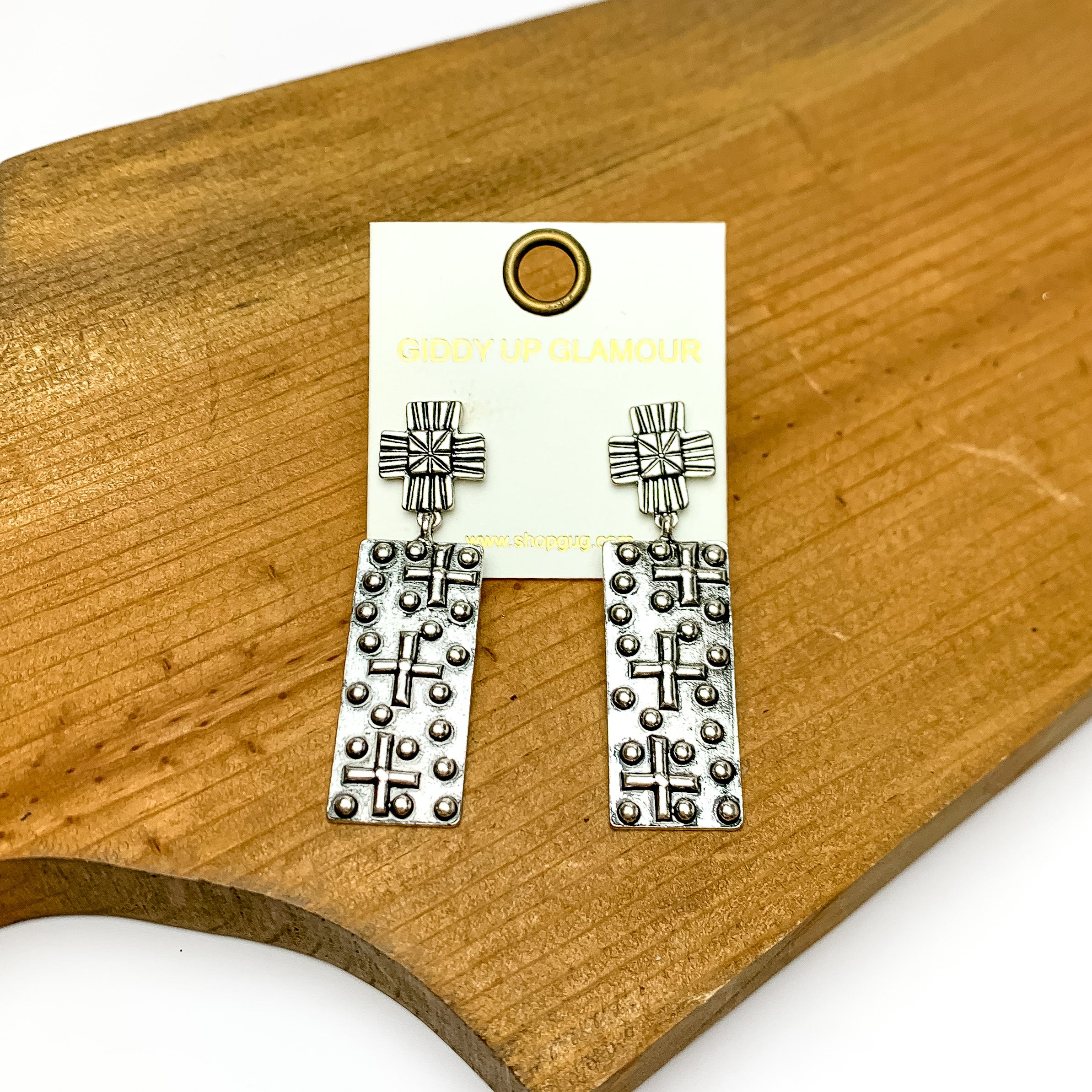 Silver tone rectangular earrings with crosses designed on them. Pictured on a piece of wood.