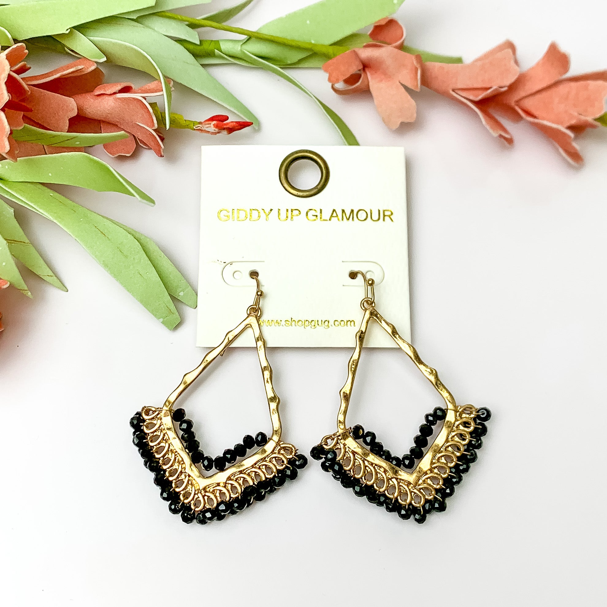 Black crystal beads bordering open drop gold tone earrings. Pictured on a white background with flowers on top.