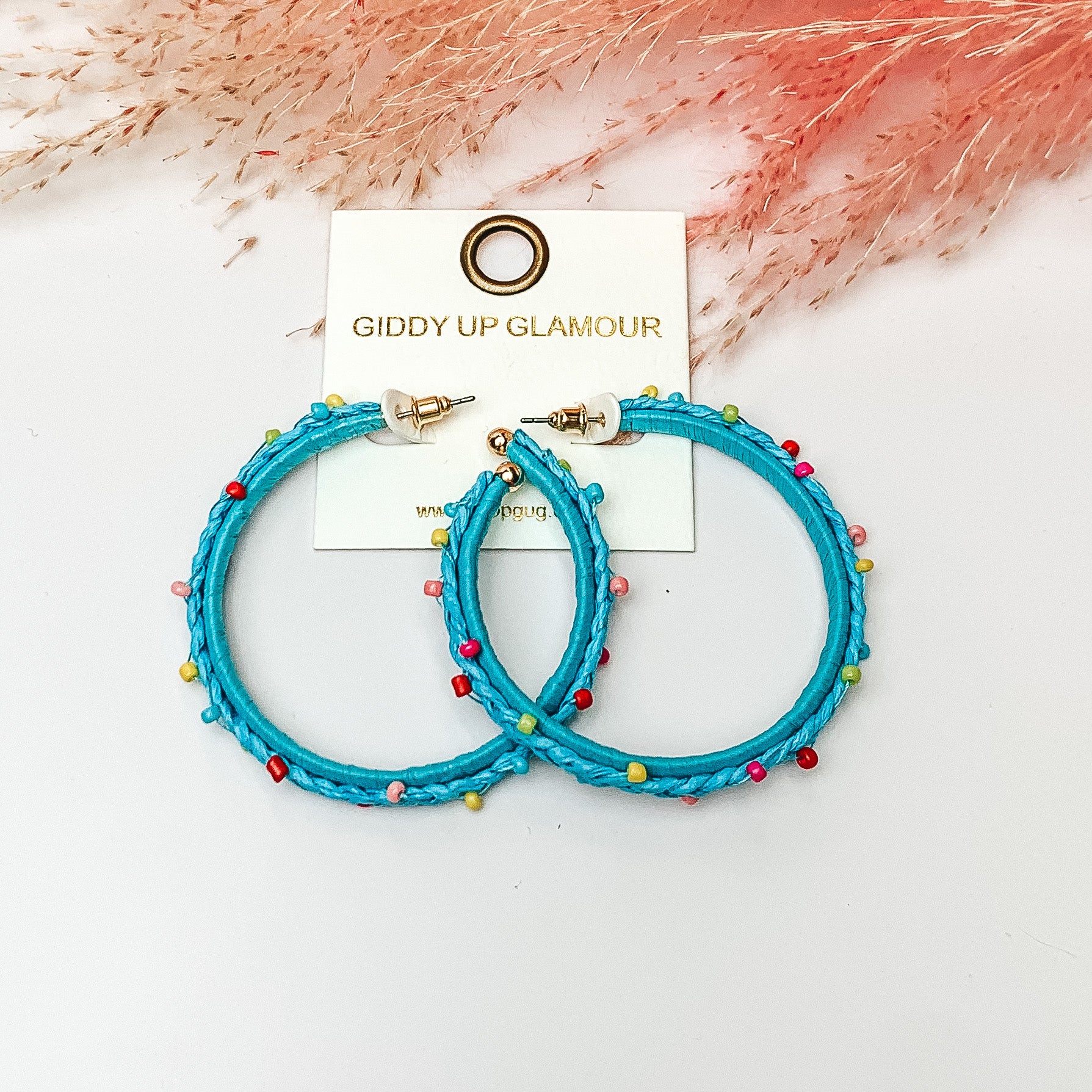 Pictured are raffia braided hoop earrings in aqua with colorful beads. They are pictured with a pink feather on a white background.