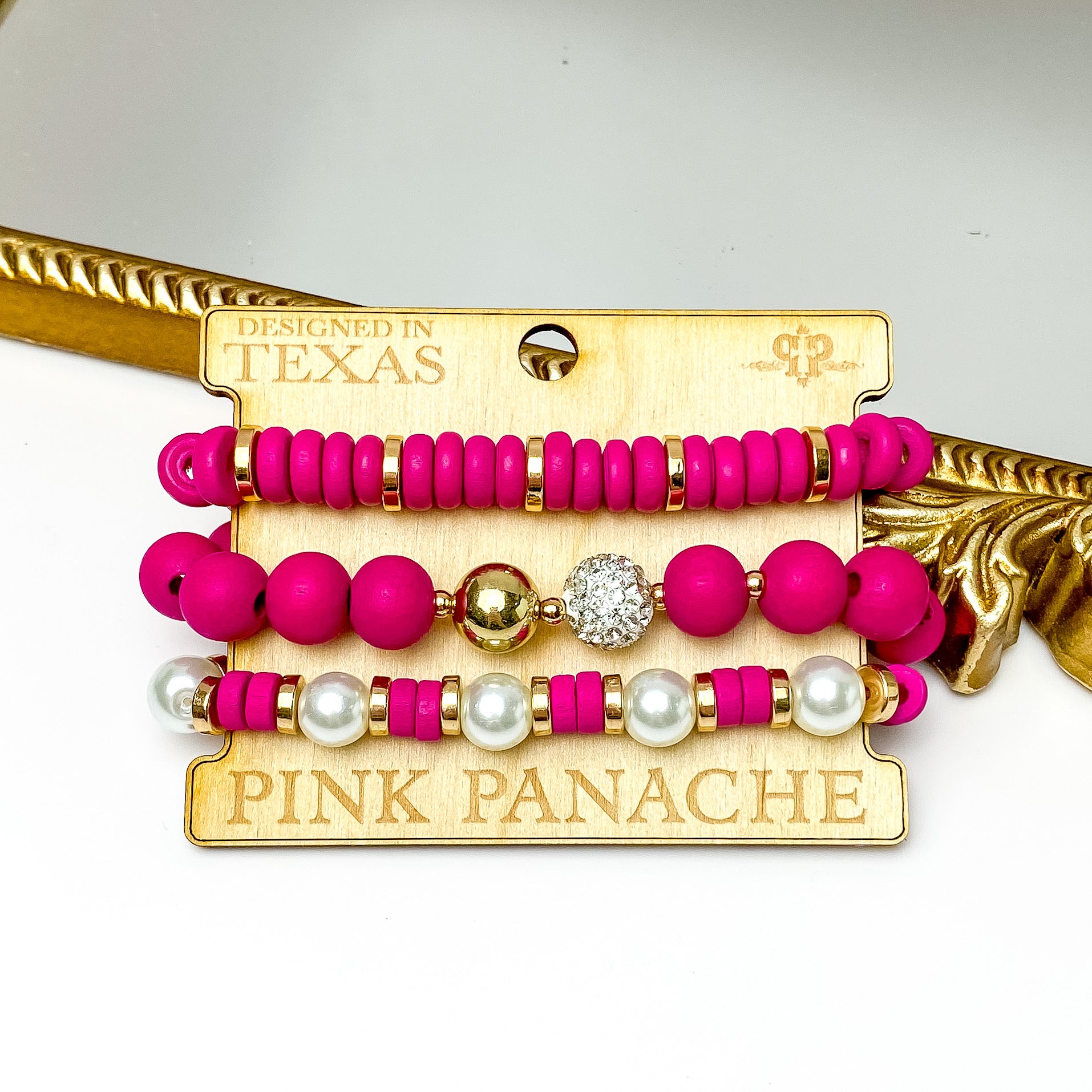 Set of three fuchsia, gold, and white pearl bead bracelets. These bracelets are pictured on a Pink Panache wood holder in front of a gold mirror and on a white background.