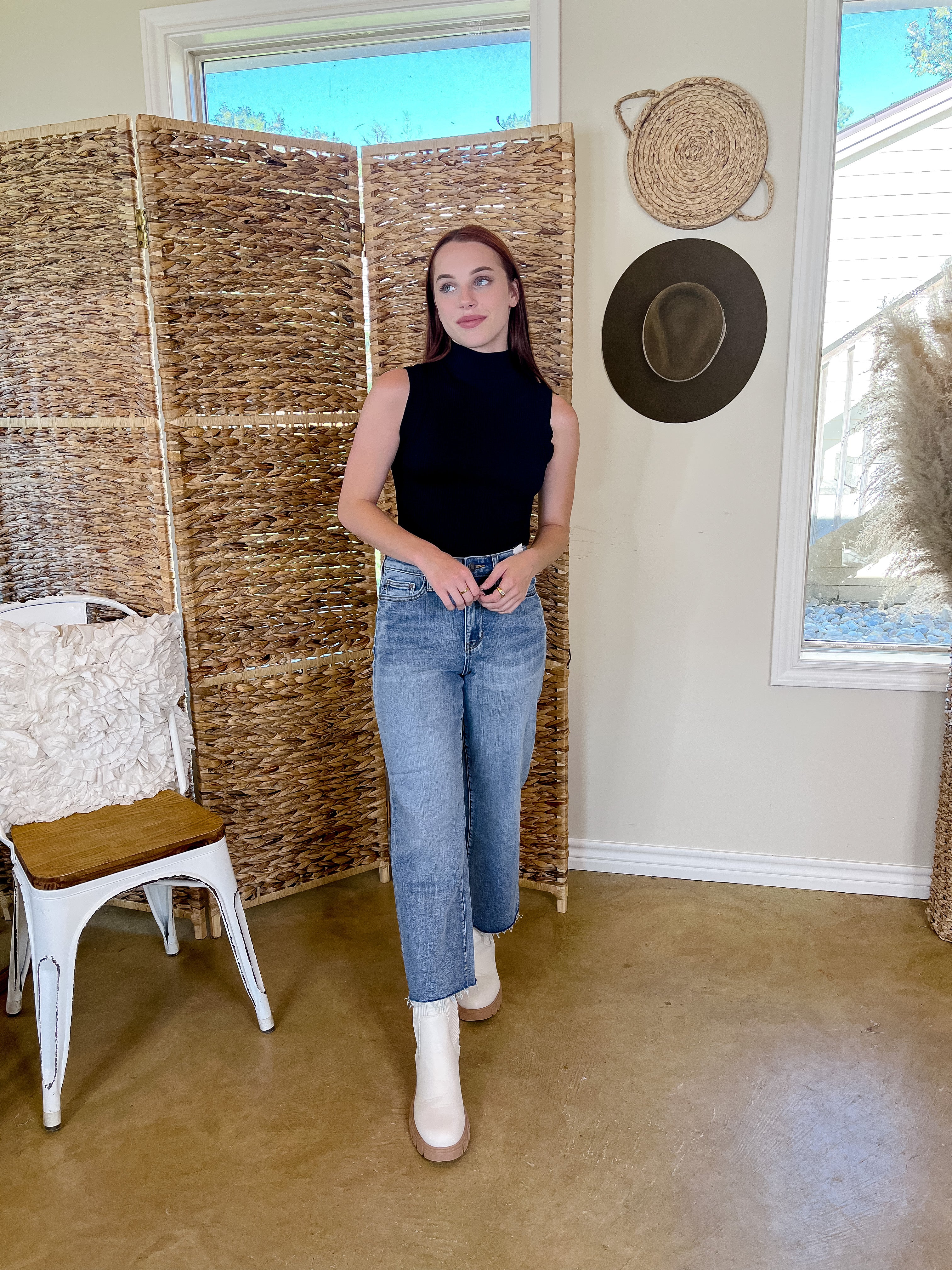 Mode is wearing a black, ribbed, turtleneck, tank top bodysuit with light wash jeans. Light wash jeans have a raw hem and she is also wearing white booties.