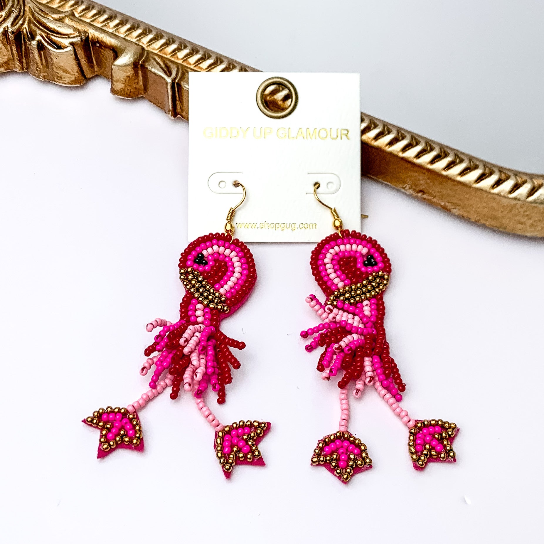These earrings include multiple color pink beads and a gold detailing. Hanging from the bottom of the flamingo shaped beaded pendant has dangling legs. These earrings are pictured leaning agasint a gold mirror on a white background.