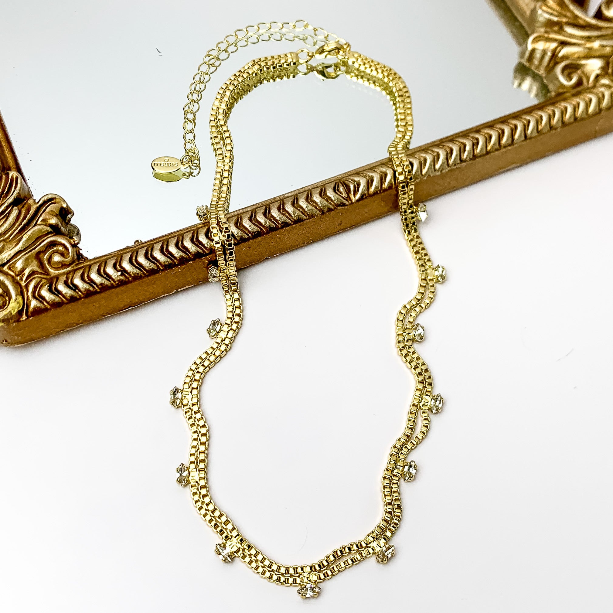 Pictured is a double, gold box chain necklace with clear crystal charm. This necklace is pictured partially laying on a gold mirror on a white background.    