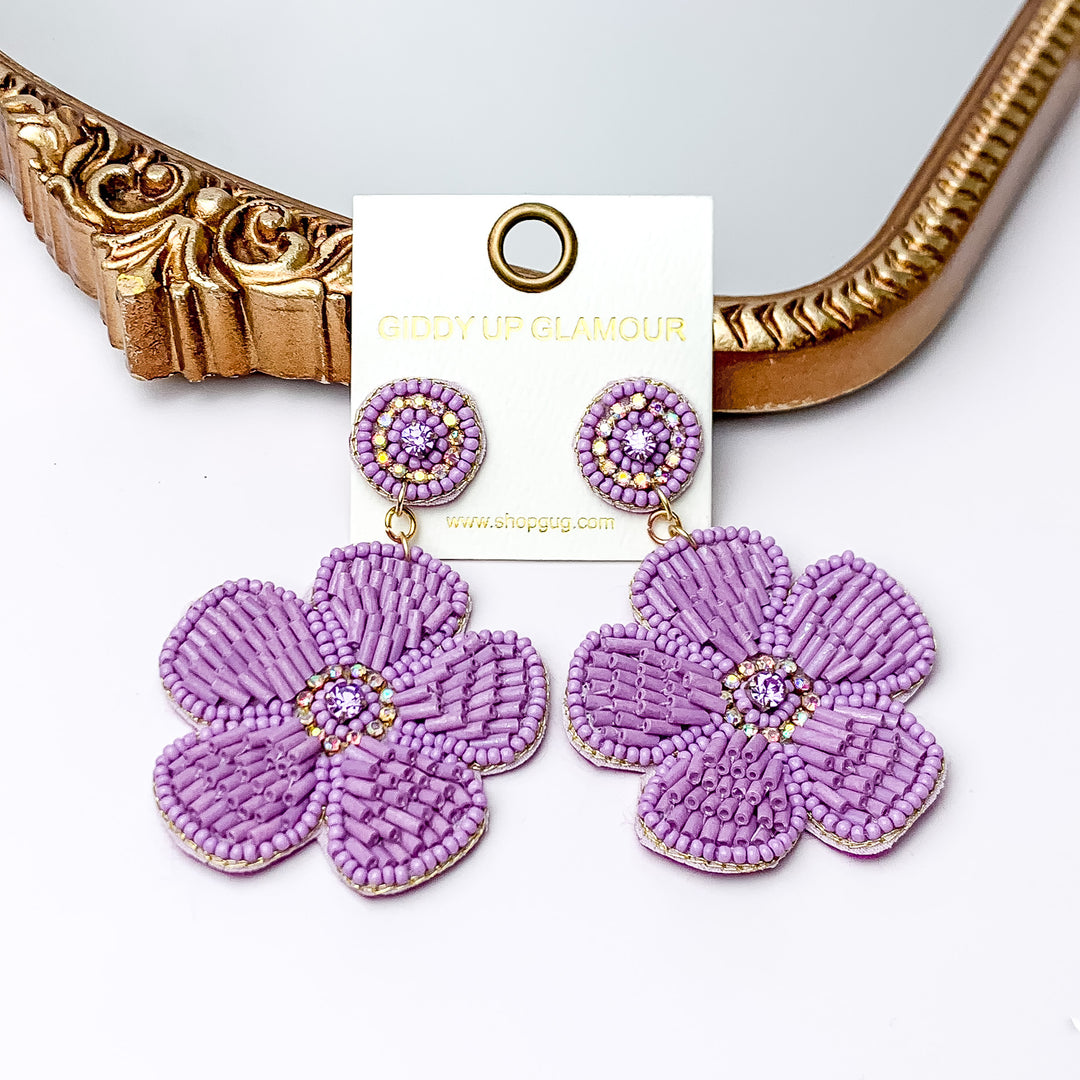 There is hanging beaded flower pendant from the gold stud earrings. The flower pendants are gold with lavender outline and detailing. These earrings are pictured in front of a gold mirror on a white background.