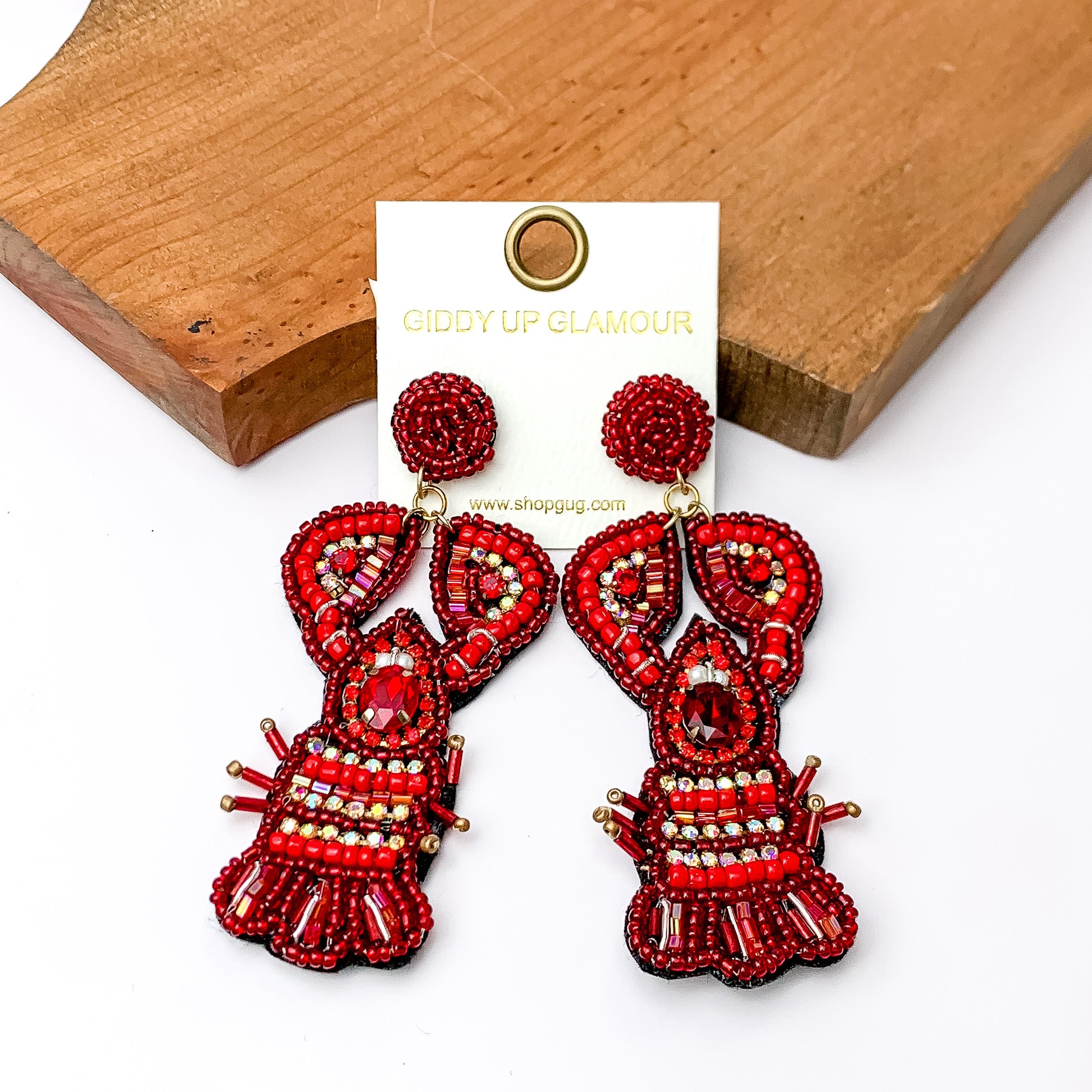 Pictured are beaded lobster earrings with red beads with ab crystal detailing. These earrings are propped up on a piece of wood with a white background.