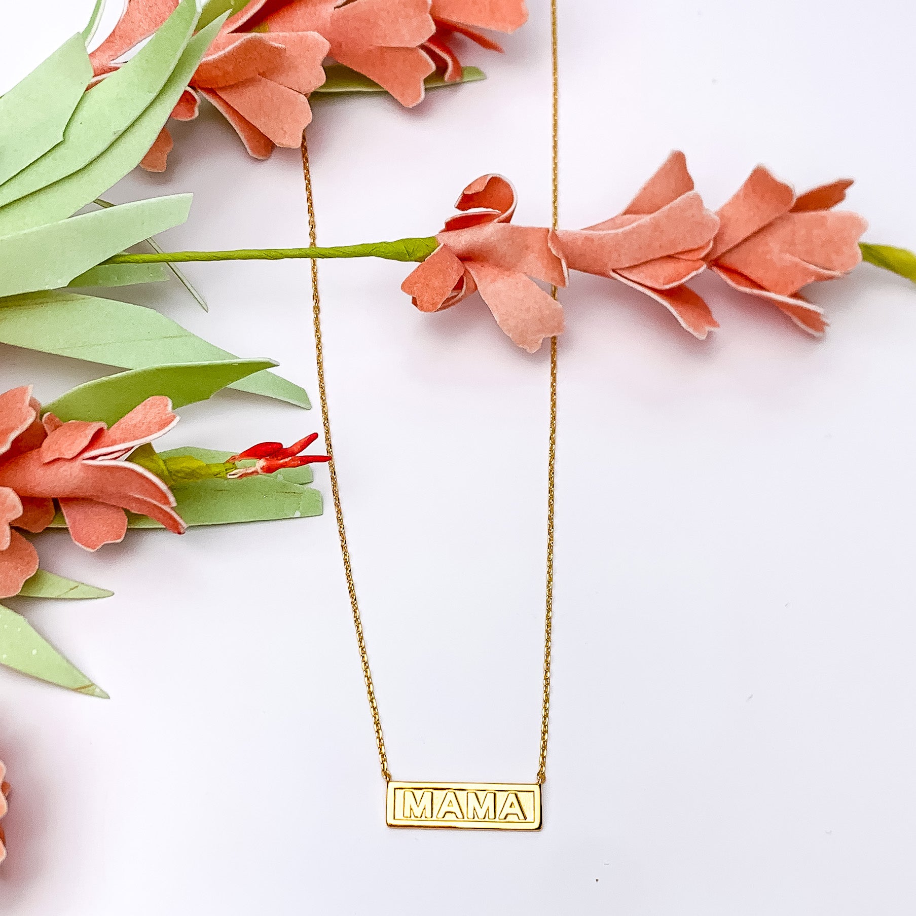 Gold tone mama plaque necklace. Pictured on a white background with flowers at the left side through the screen.