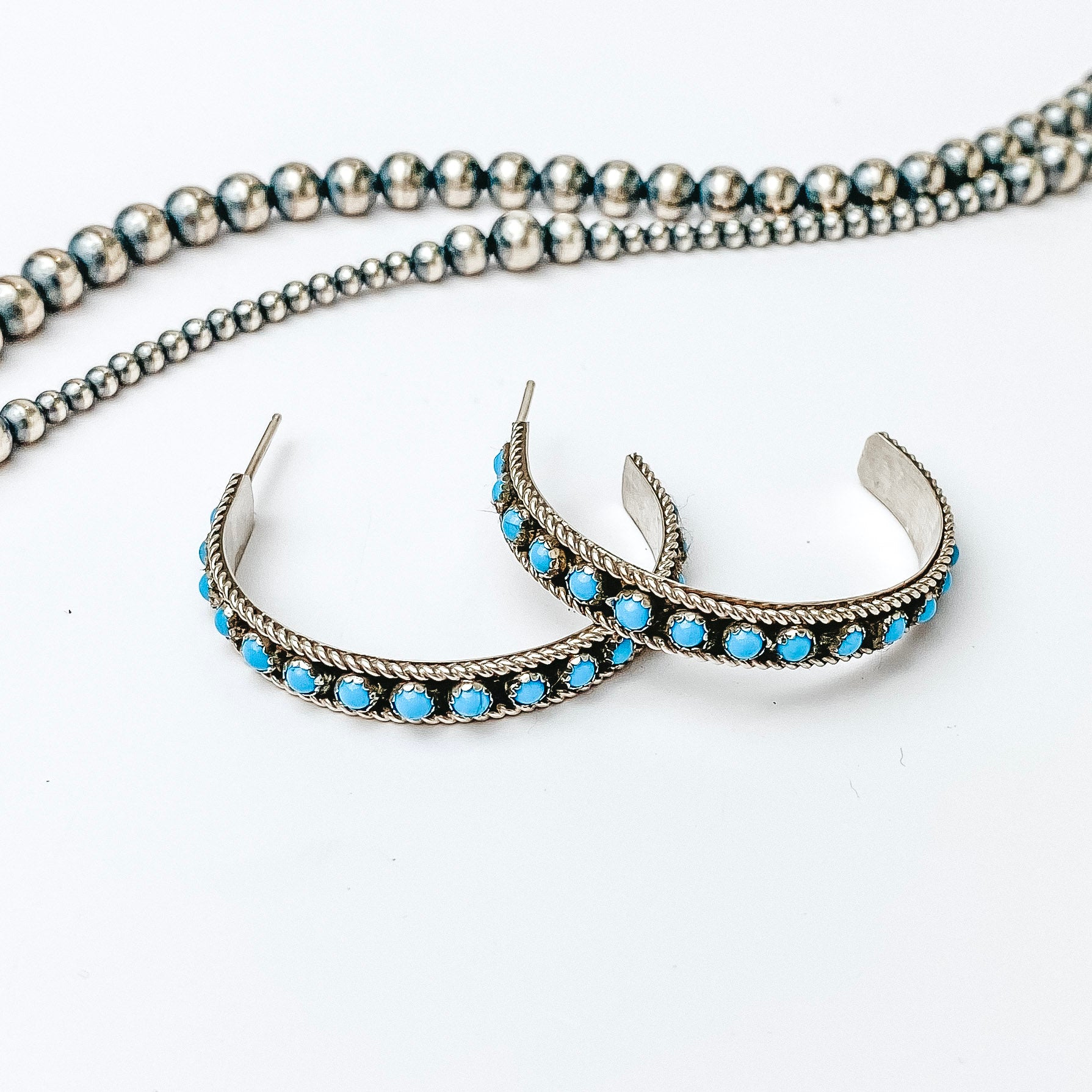 Centered is a sterling silver turquoise hoop set. Above these hoops are navajo pearls on a white background. 