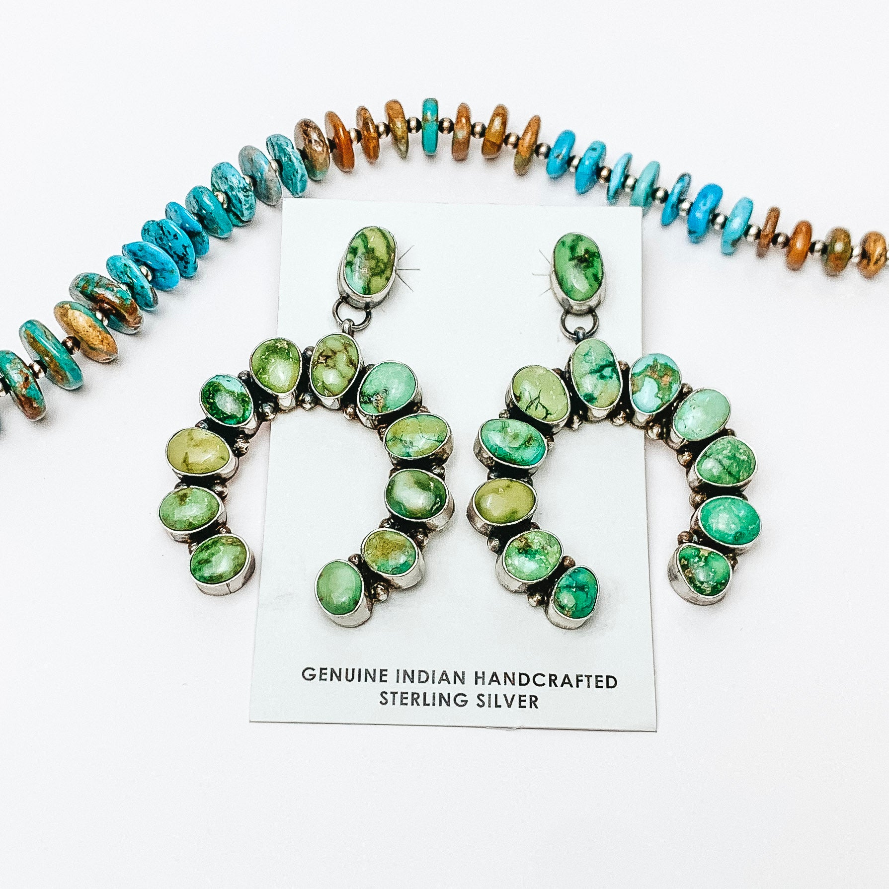 In the center of the picture is Naja shaped genuine Navajo earrings. A turquoise necklace is placed above the earrings. All on a white background. 
