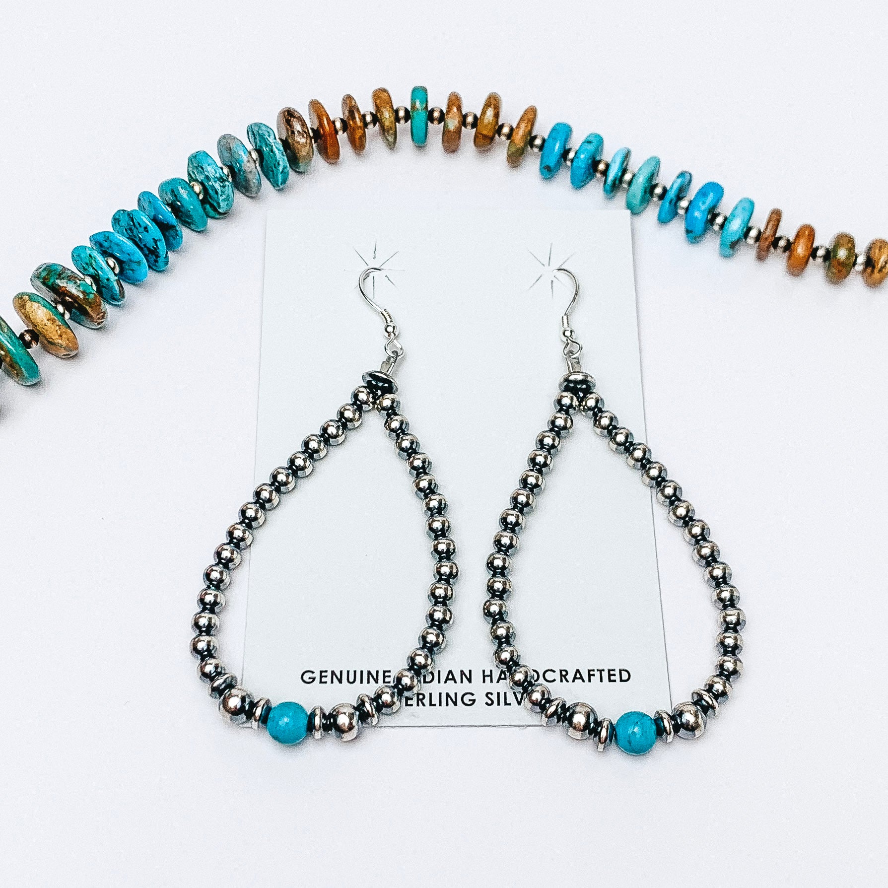 Centered in the picture is teardrop earrings strung in navajo pearls with a single turquoise stone at the bottom. A turquoise necklace sits above the earrings. Background is solid white. 