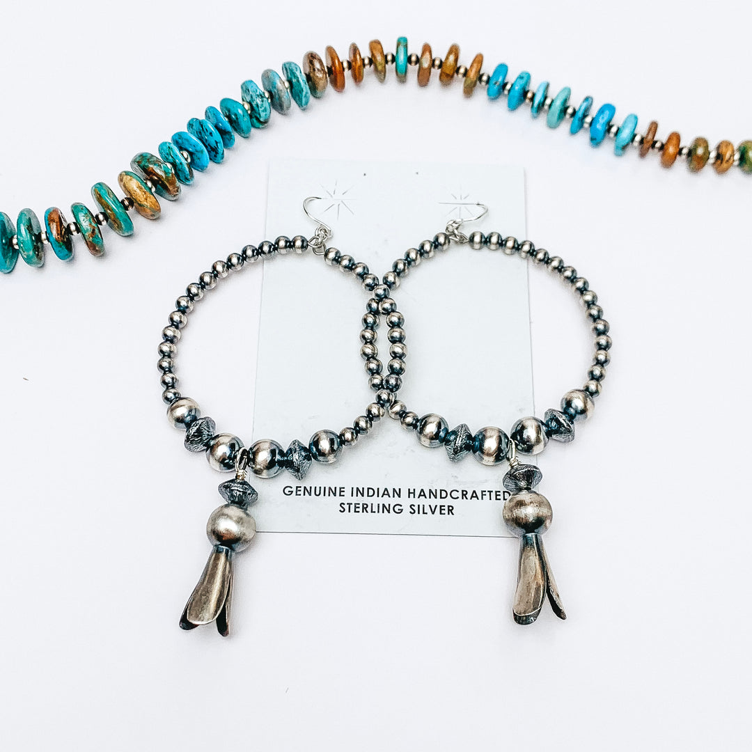 A pair of sterling silver hoop earrings made with silver sphere beads with a squash blossom pendant at the bottom of each. Pictured on a white background with genuine navajo pearls.