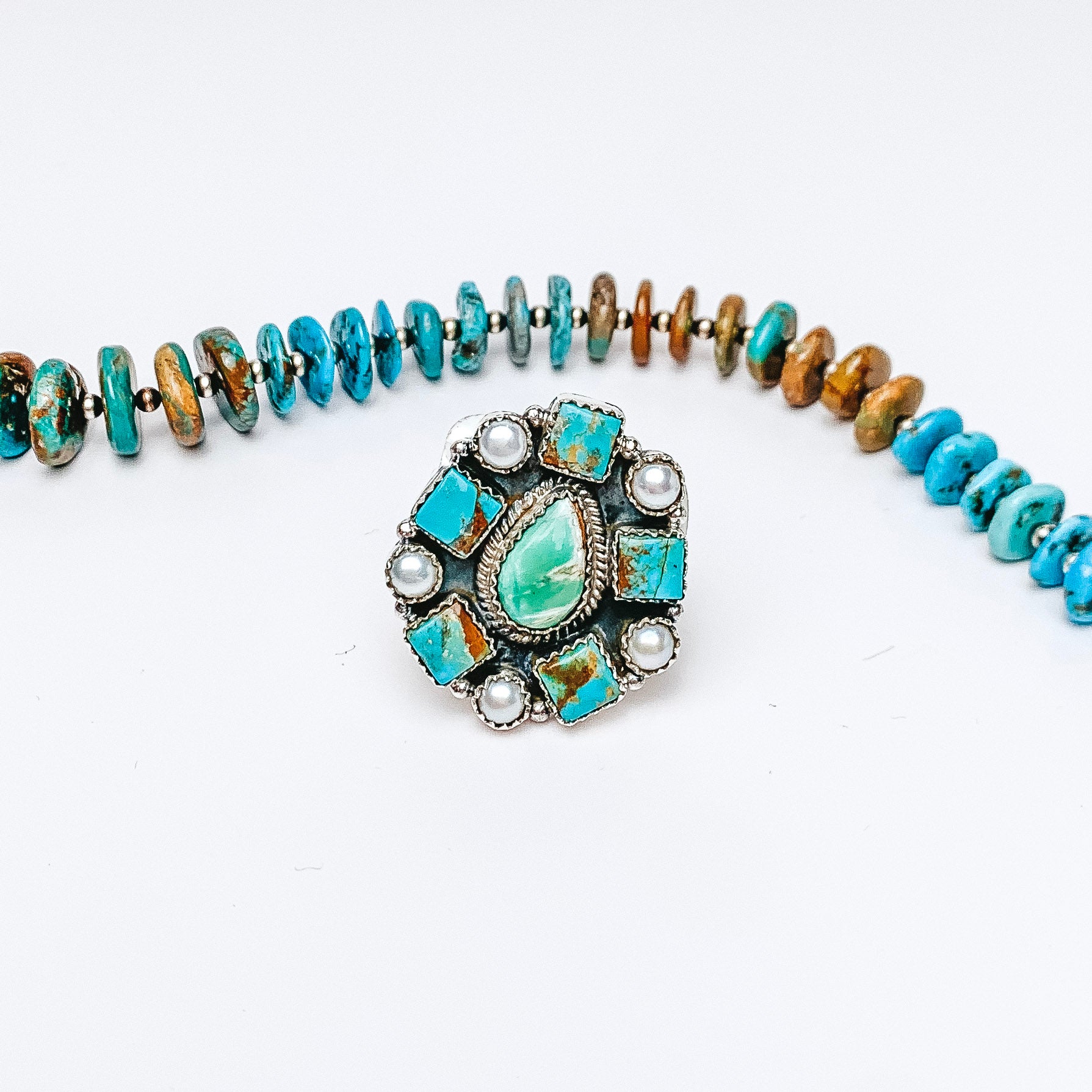 Centered in the picture is a cluster stone ring with pearl and turquoise. A turquoise necklace is above the ring. All on a white background. 