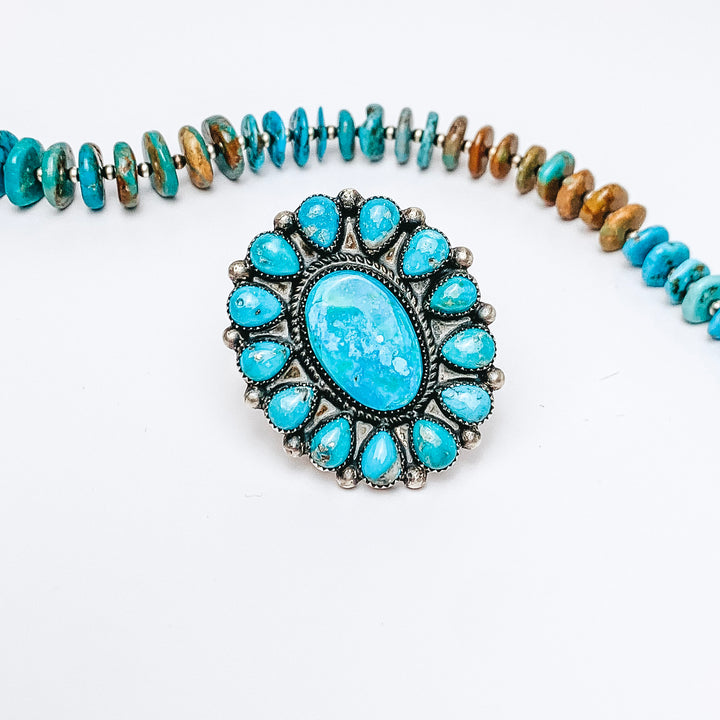 Centered is a adjustable turquoise cluster ring with a large center stone. A turquoise is laid above the ring. All on a white background. 