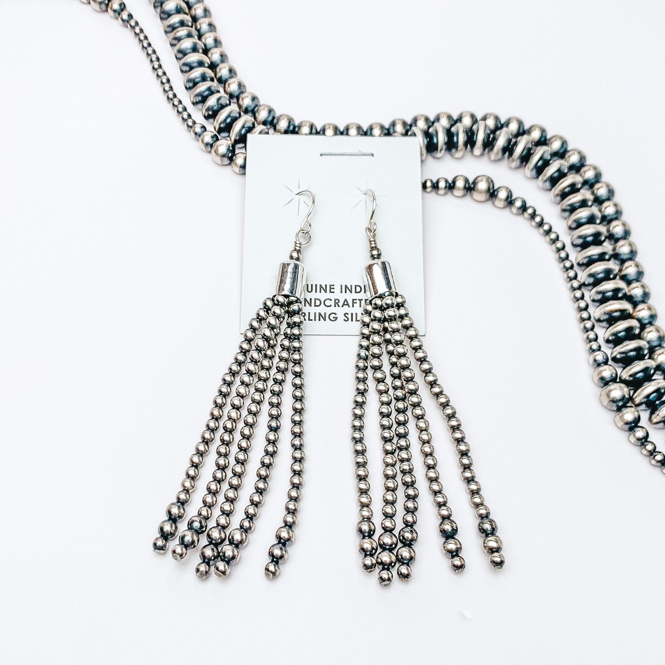 Centered in the picture is navajo pearls tassel earrings. Navajo pearls are laid above the earrings, all on a white background. 
