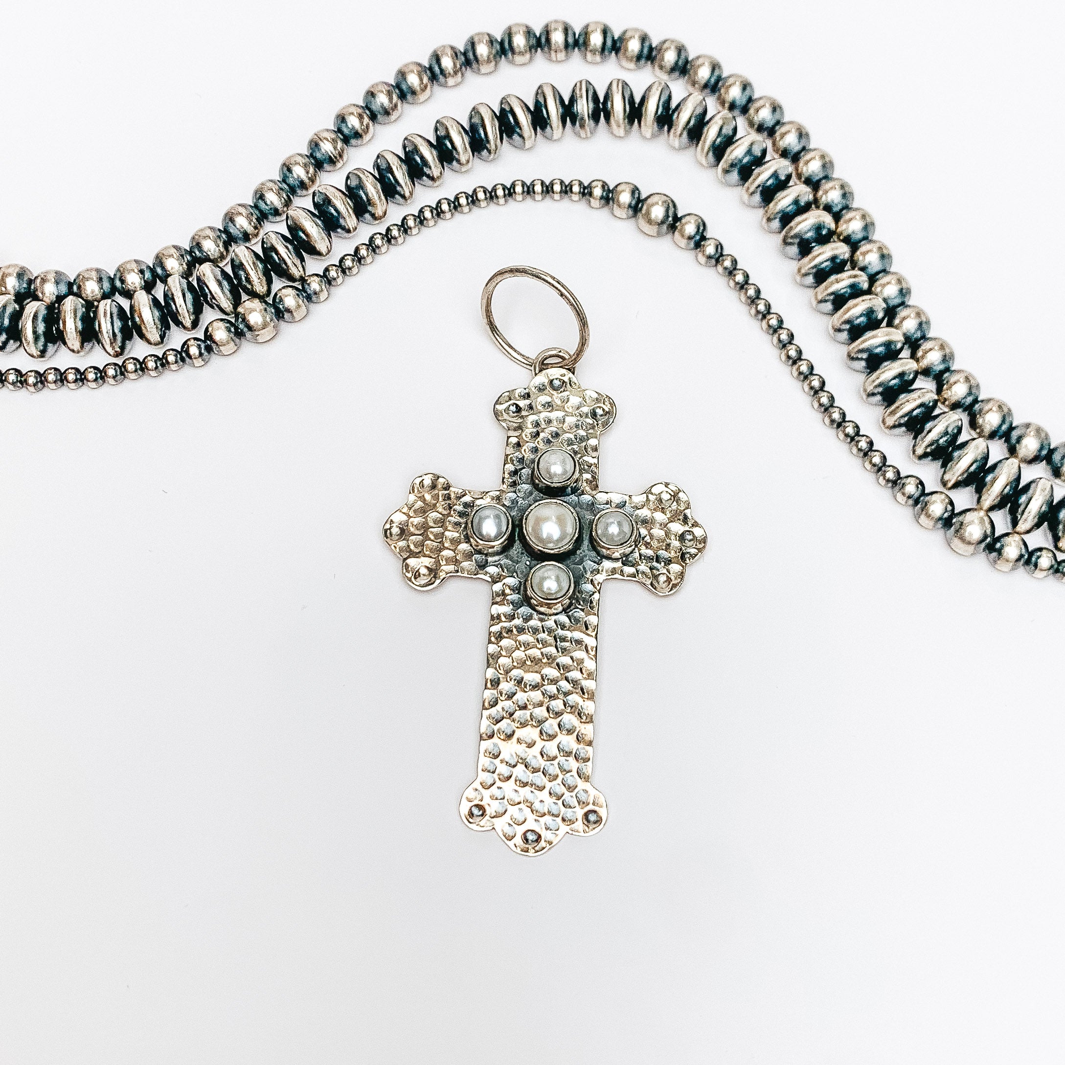 Centered in the picture is a hammered pattern cross pendant with 5 mother of pearl stones. Navajo pearls are laid above the cross, all on a white background. 