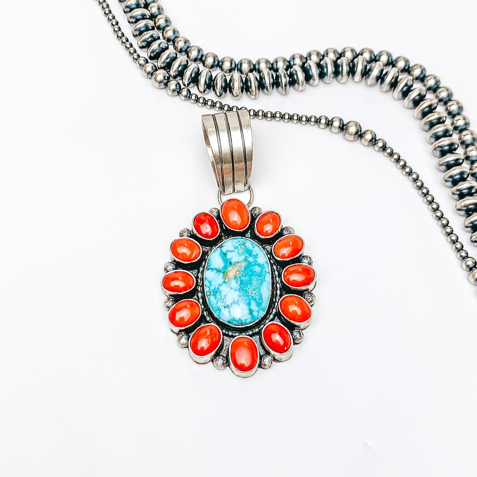 Centered in the picture is a large, round, turquoise and coral pendant. Above the pendant is navajo pearls, all on a white background. 