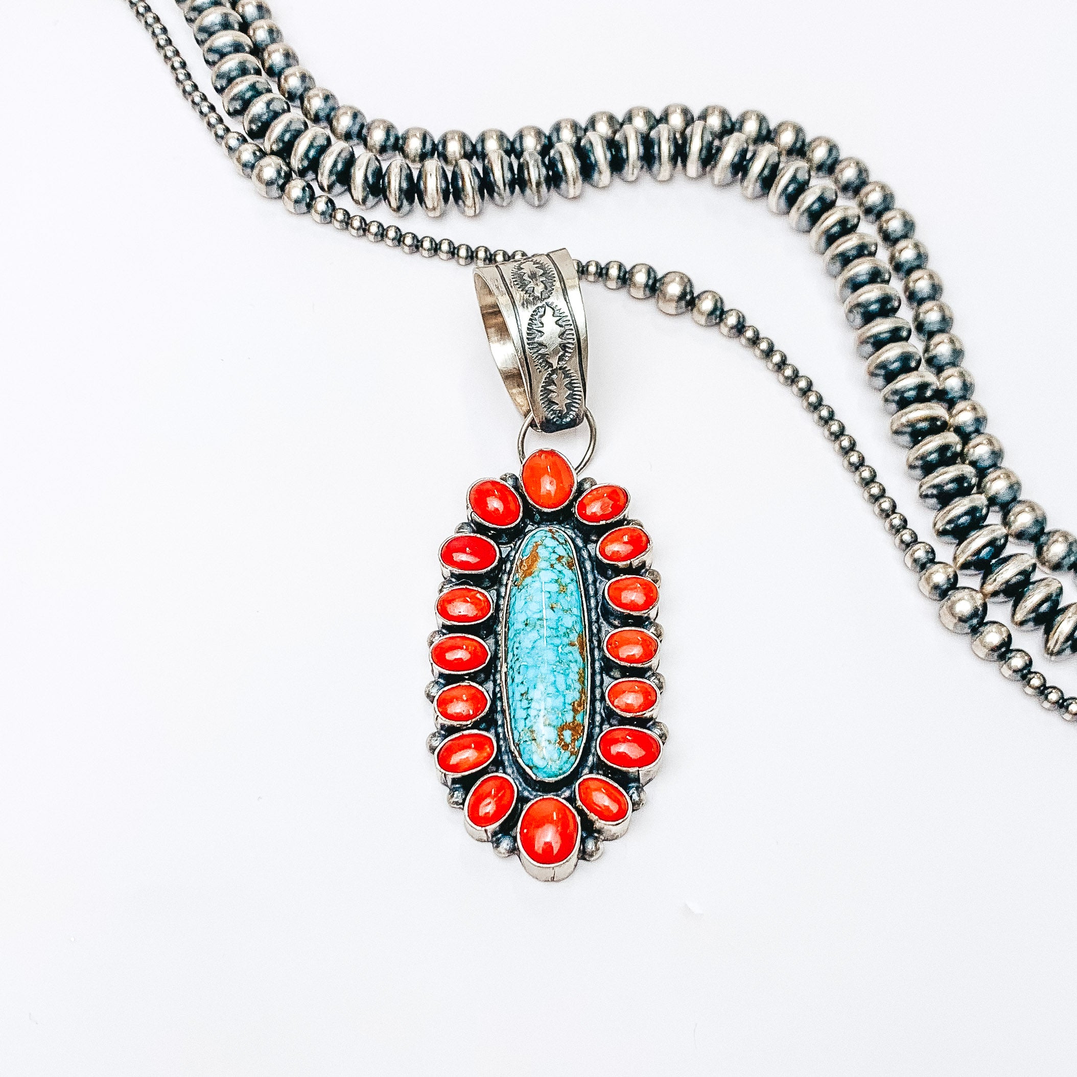 Centered in the picture is a large, oval, turquoise and coral pendant. Above the pendant is navajo pearls, all on a white background.