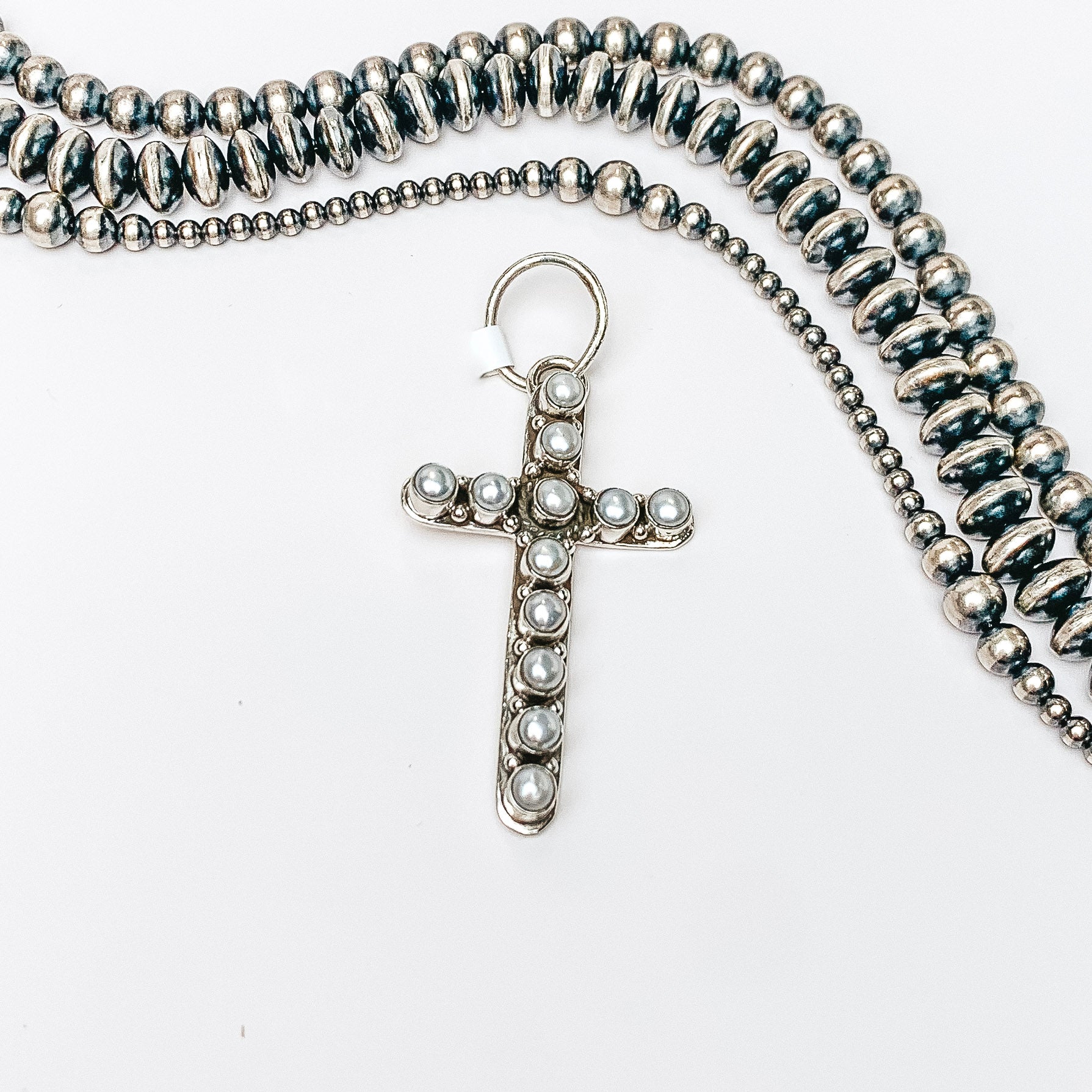 Centered in the picture is a small cross pendant with mother of pearl stones. Above the cross is navajo pearls, all on a white background. 