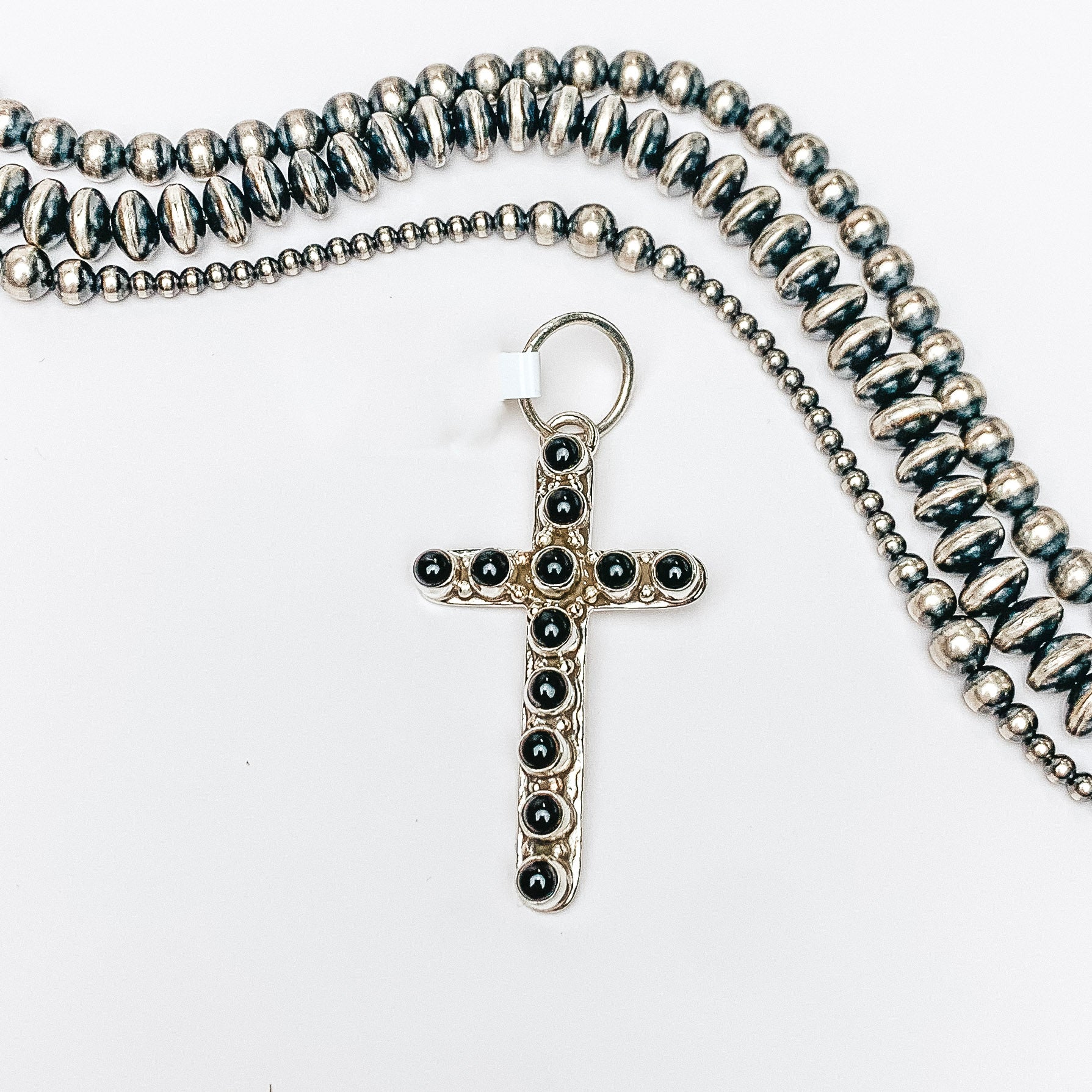Centered in the picture is a small cross pendant with black onyx stones. Navajo pearls are laid to the top of the pendant. ALl on a white background. 