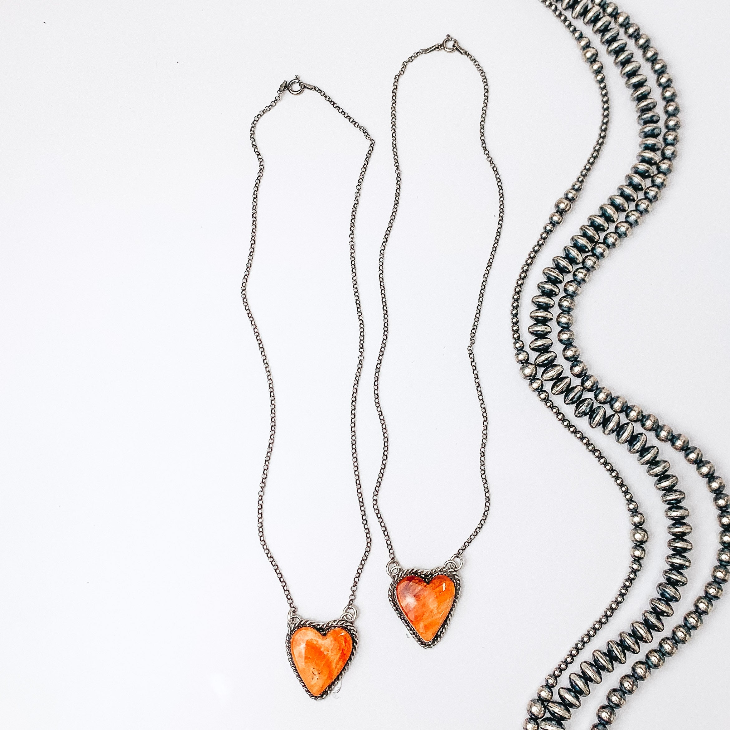 Centered in the middle of the picture is a silver necklace with a spiny oyster heart pendant. Navajo pearls are laid to the right of the necklace, all on a white background. 