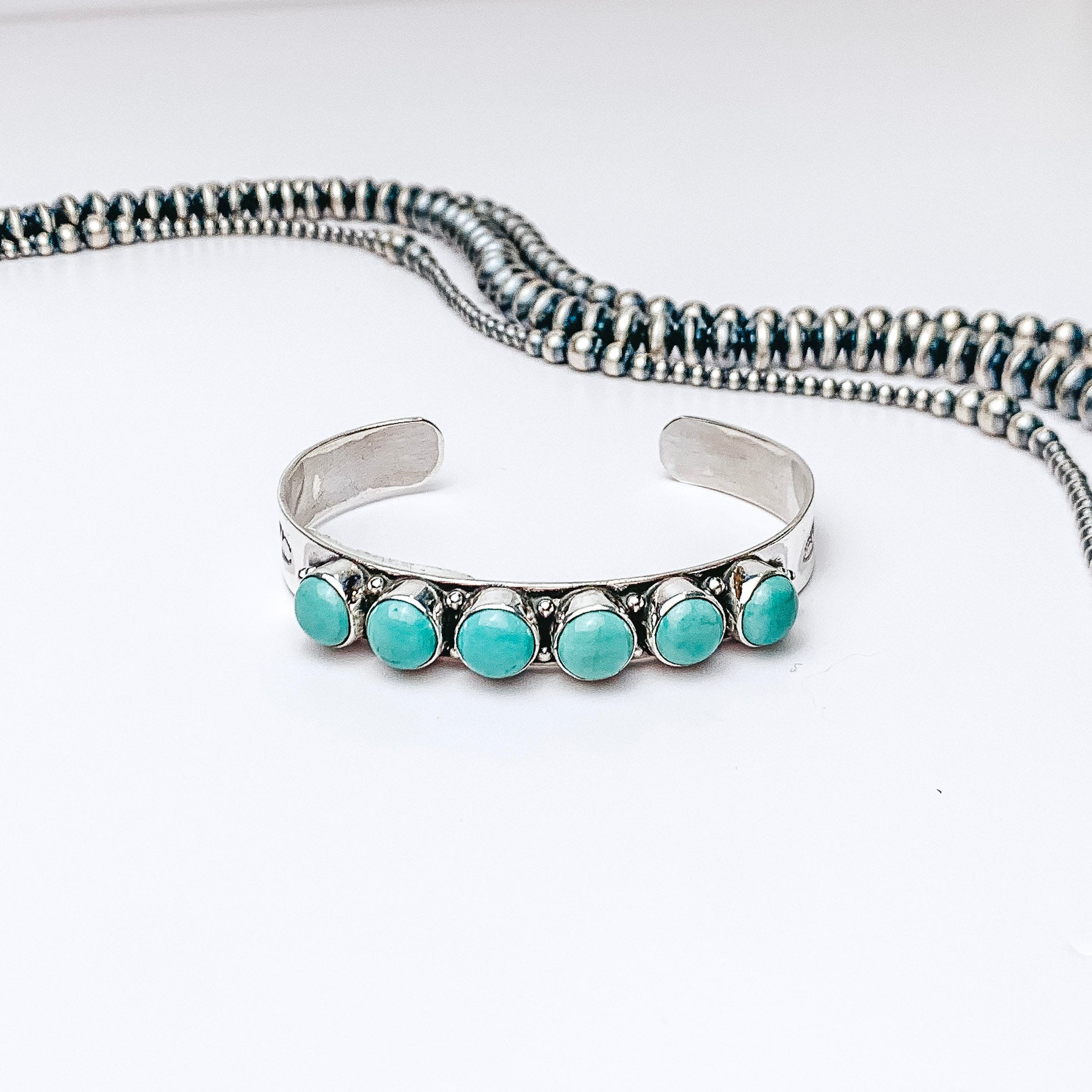 Centered in the picture is a cuff with six kingman turquoise stones. Navajo pearls are laid above the cuff, all on a white background. 