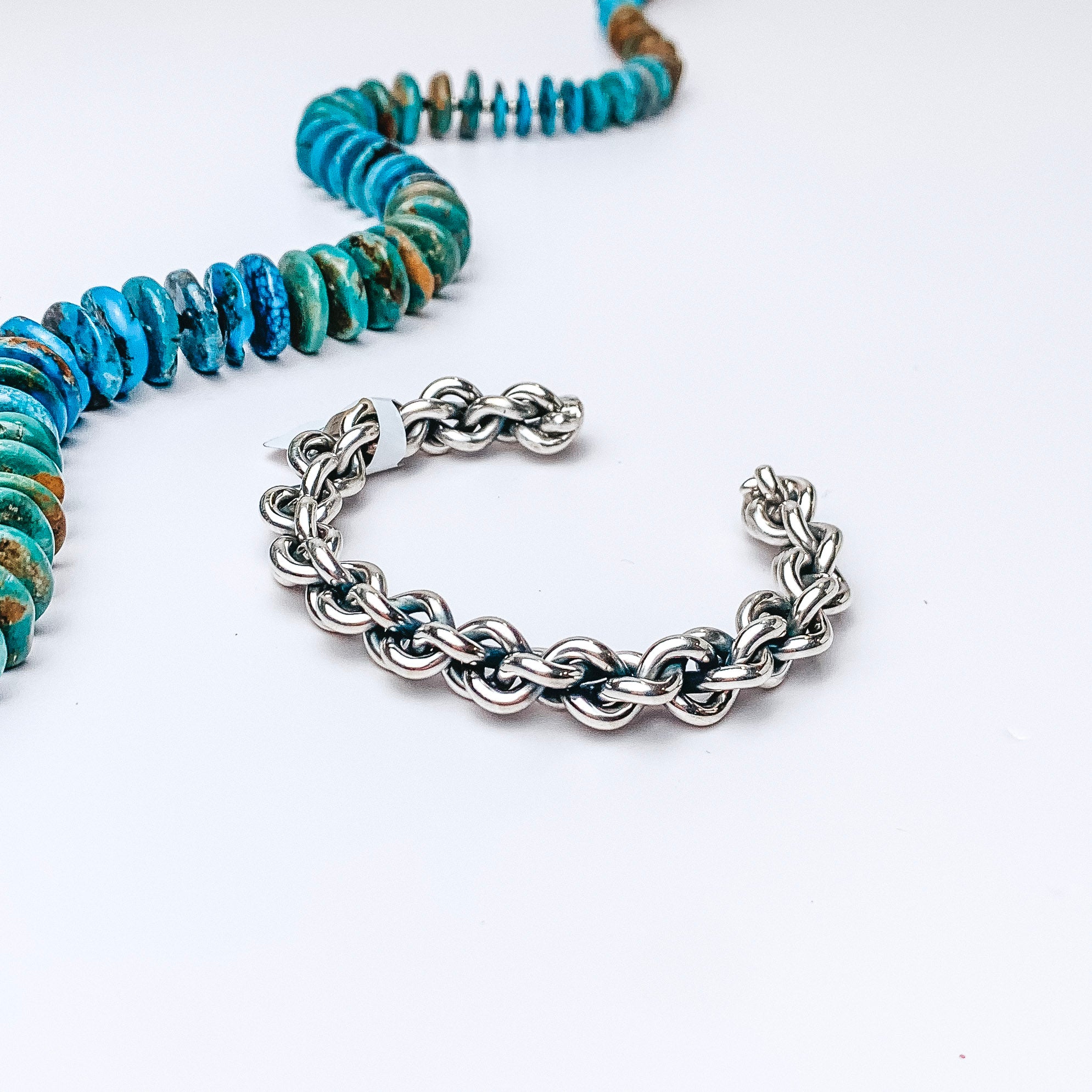 Centered in the picture is a sterling silver braided cuff. A turquoise necklace is laid above the cuff, all on a white background. 