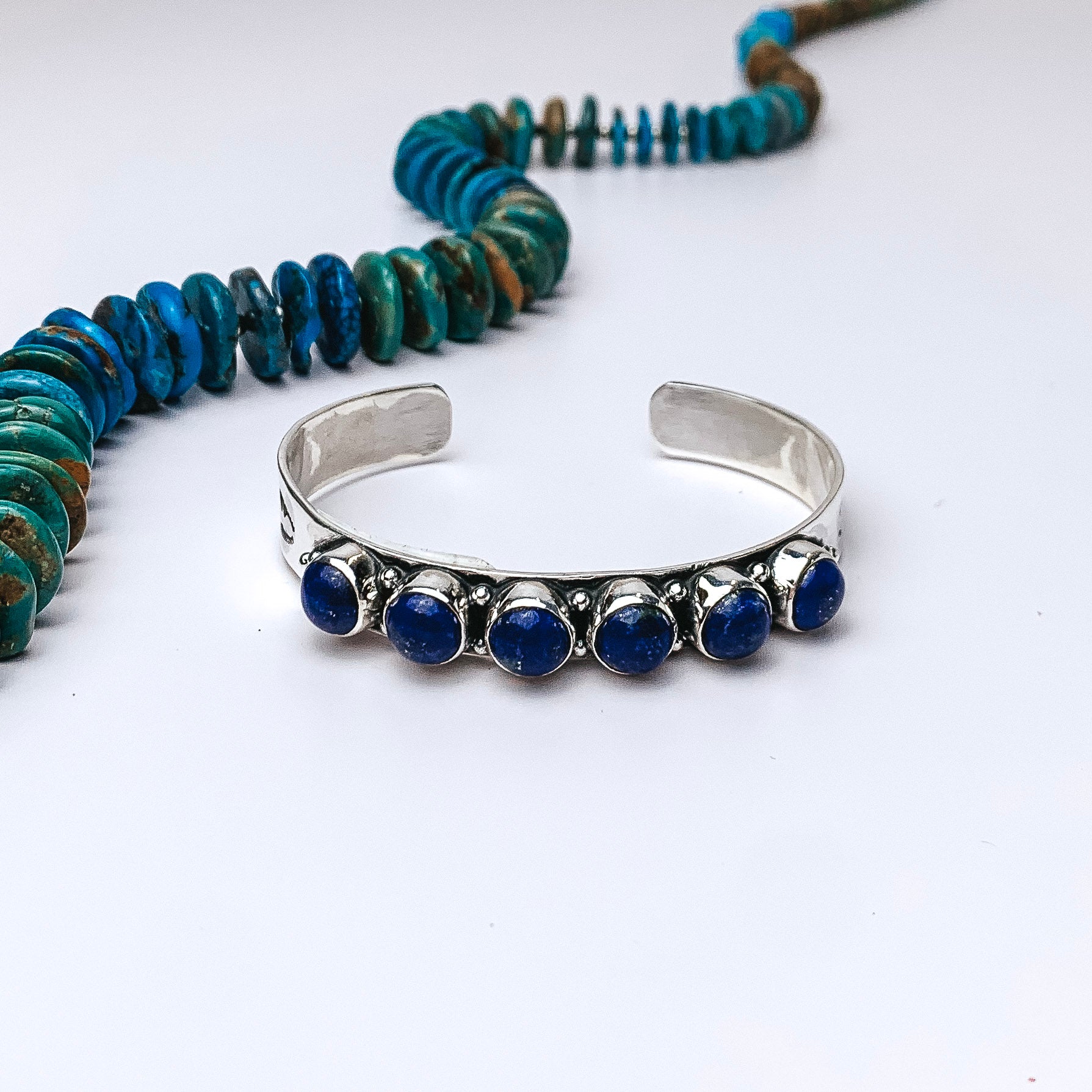 Centered in the picture is a sterling silver cuff with six dark lapis stones. Above the cuff is a turquoise necklace. All on a white background. 