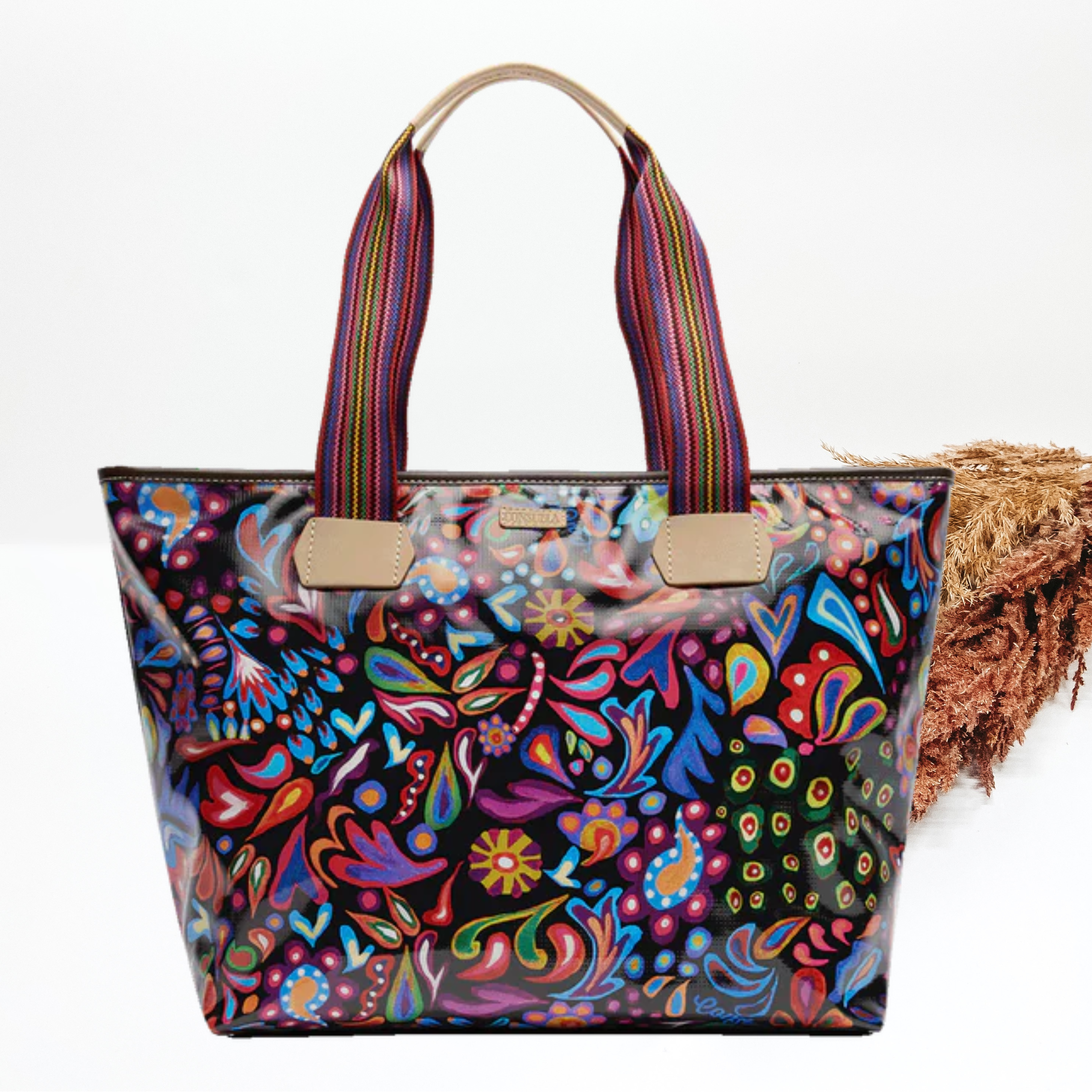 This bag is a large tote with leather handles. This tote is black with a multicolored paisley print all over. This tote is pictured in front of brown pompous grass on a white background. 