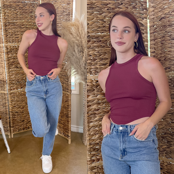 Model is wearings a maroon, scoop neck cropped tank top with light blue jeans. She is also wearing gold hoops and white tennis shoes.