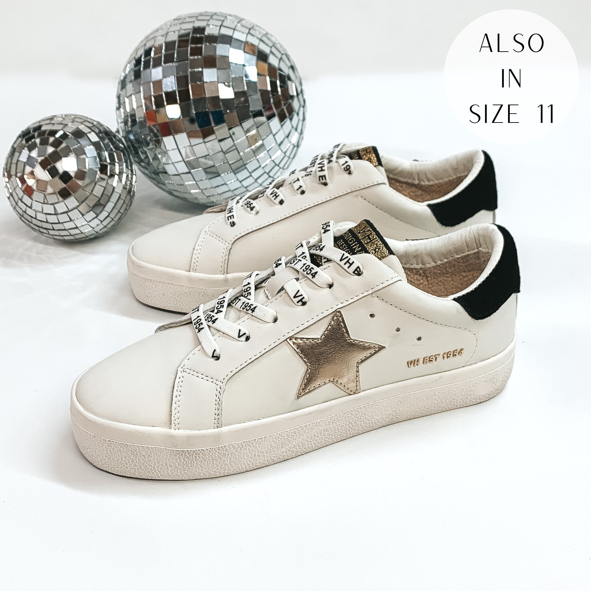 Pictured are a white pair of sneakers with a gold star emblem and a black part on the heel. These shoes are pictured on a white background with disco balls in the background.