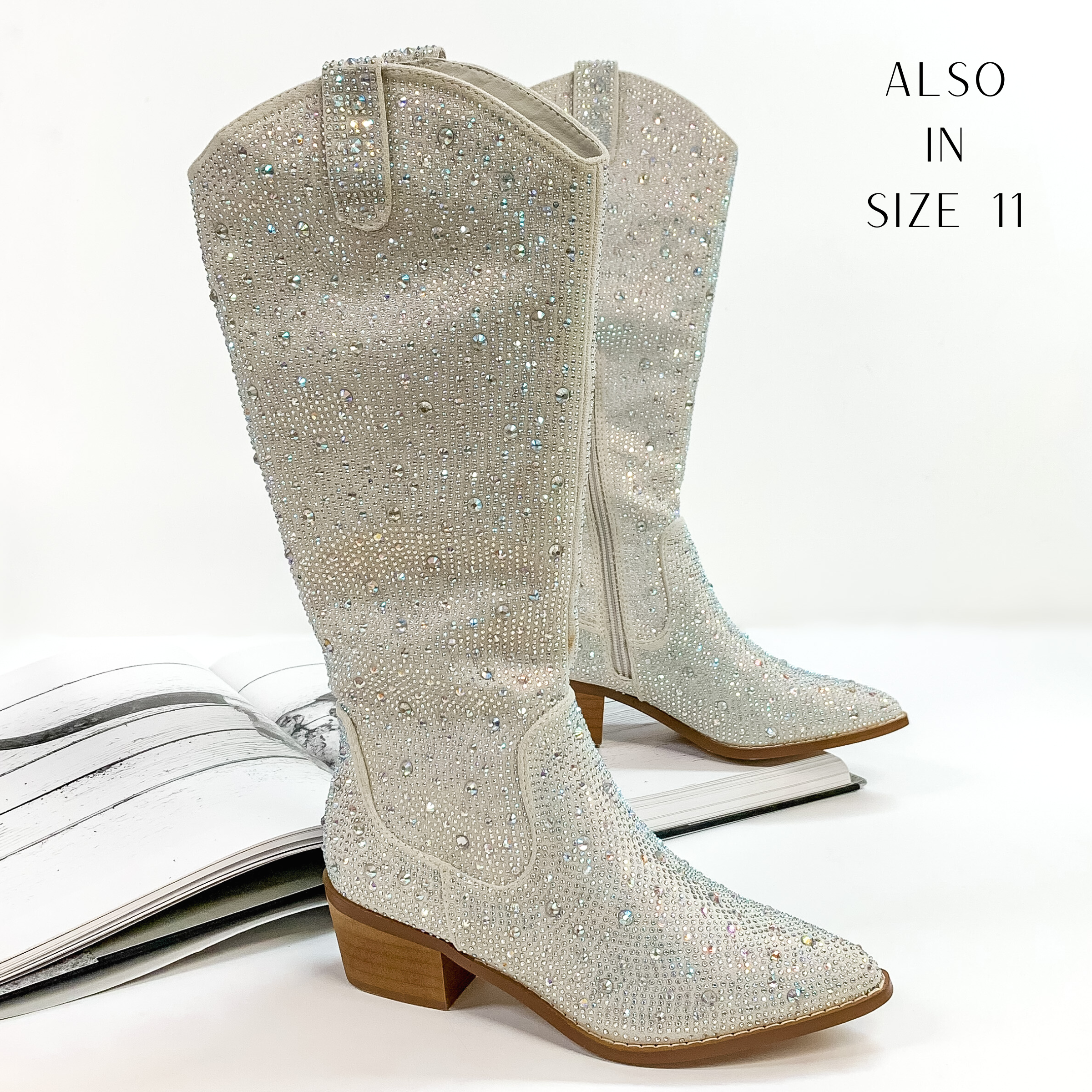 Pictured are silver cowboy boots with ab crystals covering the entire boot. These boots also include a tan heel and sole. These boots are pictured on front on a white background with one boot resting on an open book.
