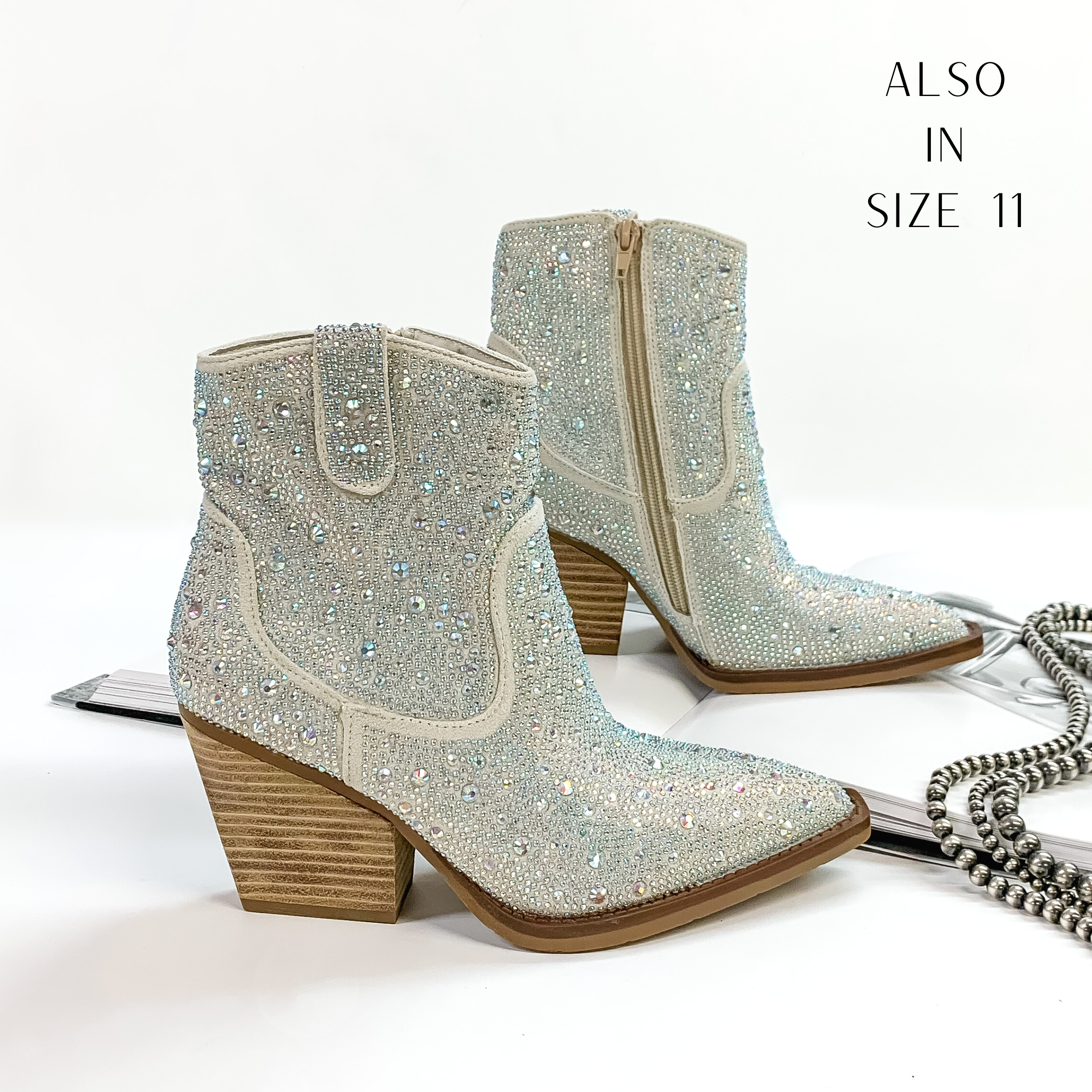 Pictured are silver cowboy booties with ab crystals covering the entire boot. These boots also include a tan heel and sole. These boots are pictured on front on a white background with one boot resting on an open book.
