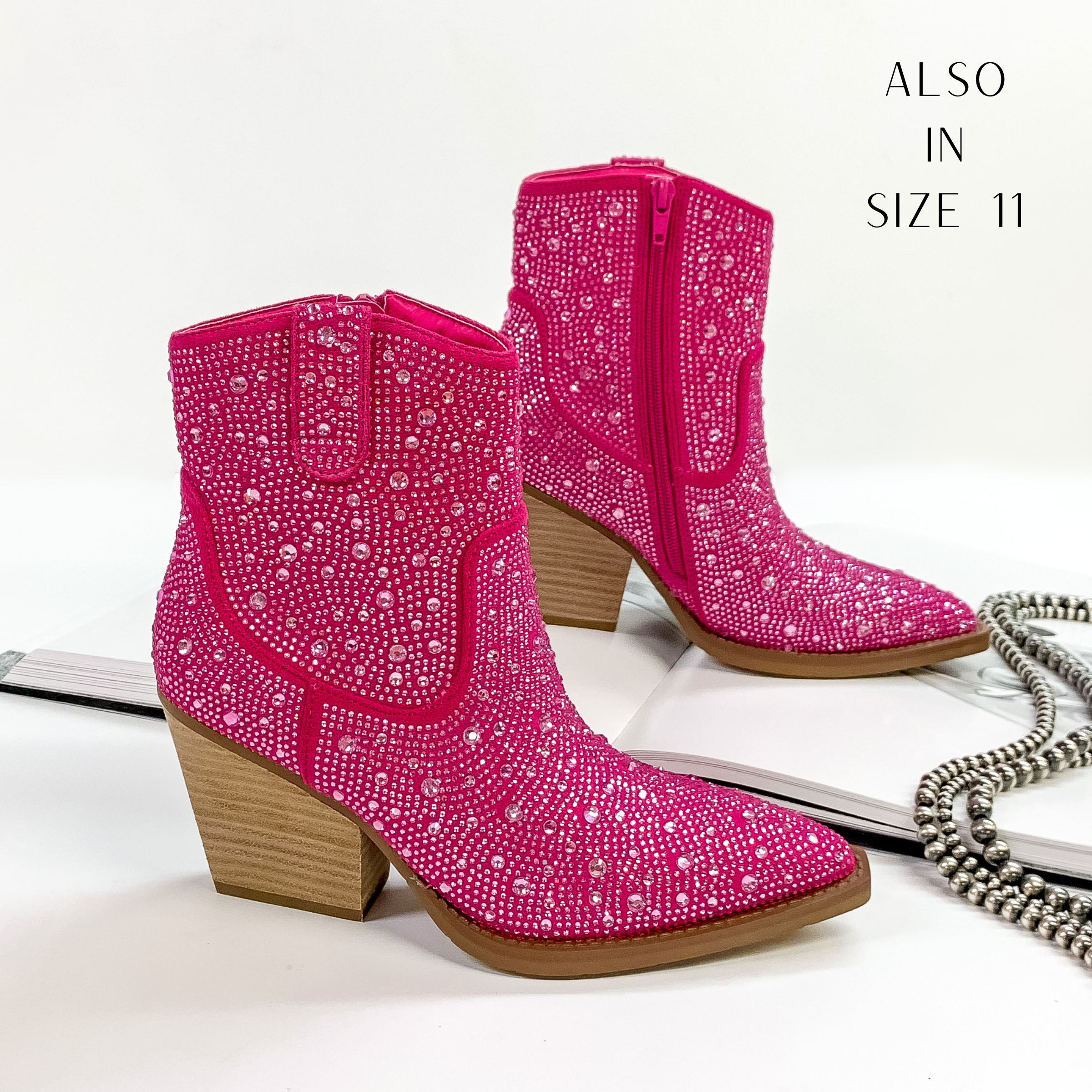 Pictured are black cowboy booties with pink crystals covering the entire boot. These boots also include a tan heel and sole. These boots are pictured on front on a white background with one boot resting on an open book.