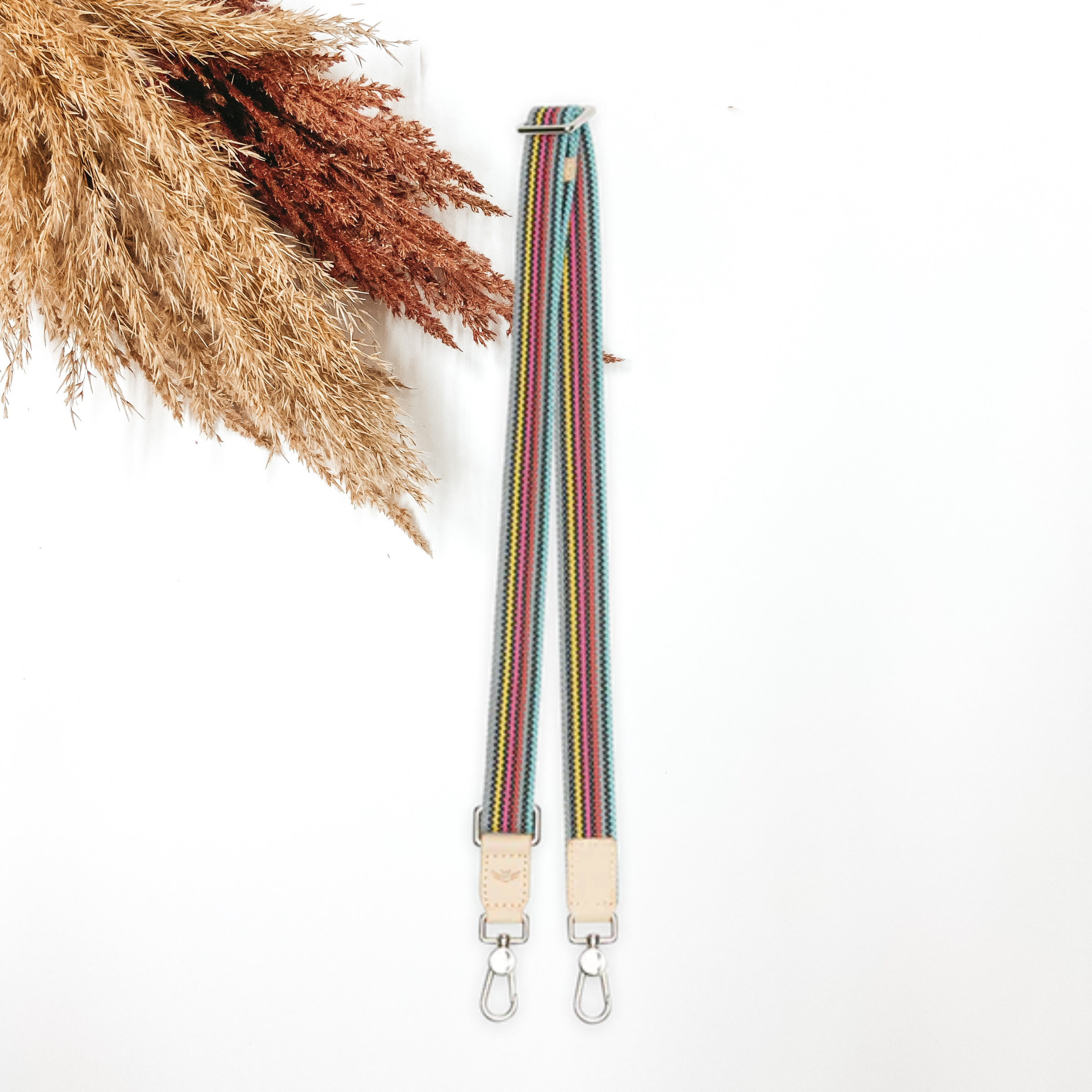 A woven multicolored purse strap that has a main color of grey. This purse strap is pictured on a white background with tan and brown pompous grass in the top left corner.