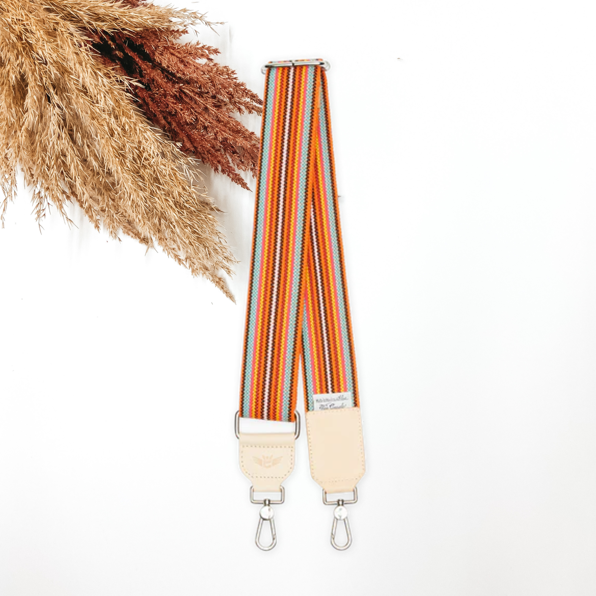 A woven multicolored striped purse strap that has a main color of orange. This purse strap is pictured on a white background with tan and brown pompous grass in the top left corner.