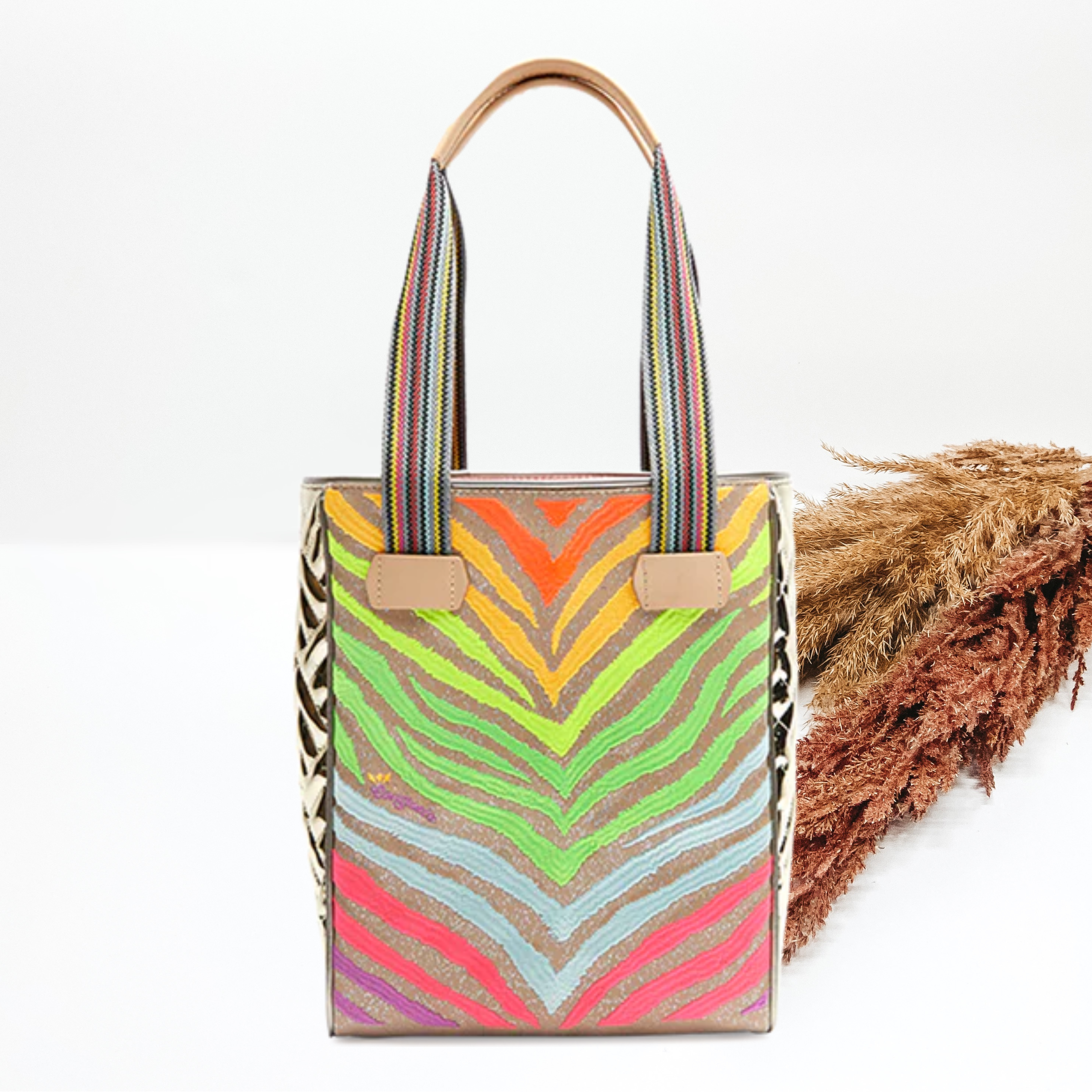 Gold tote bag with a multicolored embroidered zebra print design. The straps are striped with light tan accents. This tote is pictured on a white background with tan and brown pompous grass on the right side.