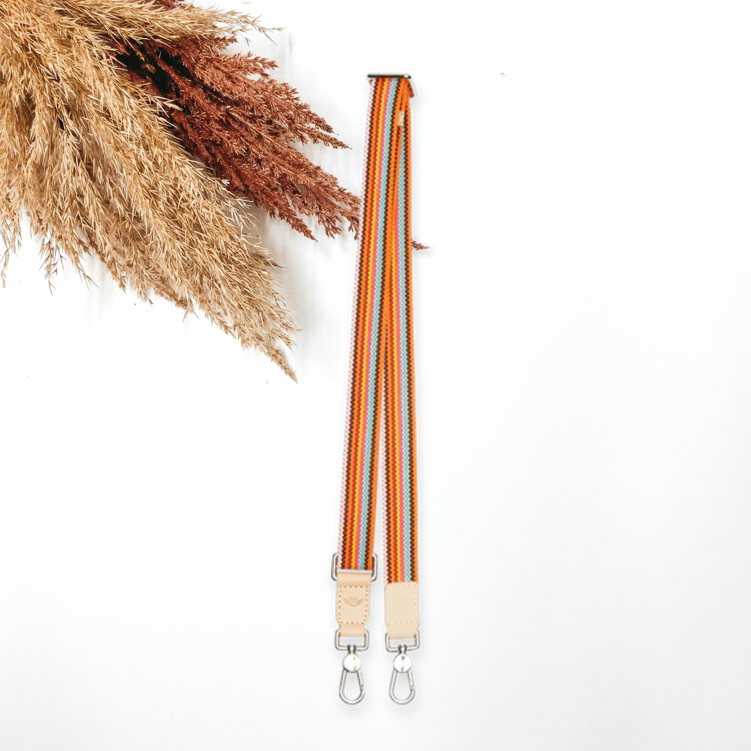 A woven multicolored purse strap that has a main color of orange. This purse strap is pictured on a white background with tan and brown pompous grass in the top left corner.