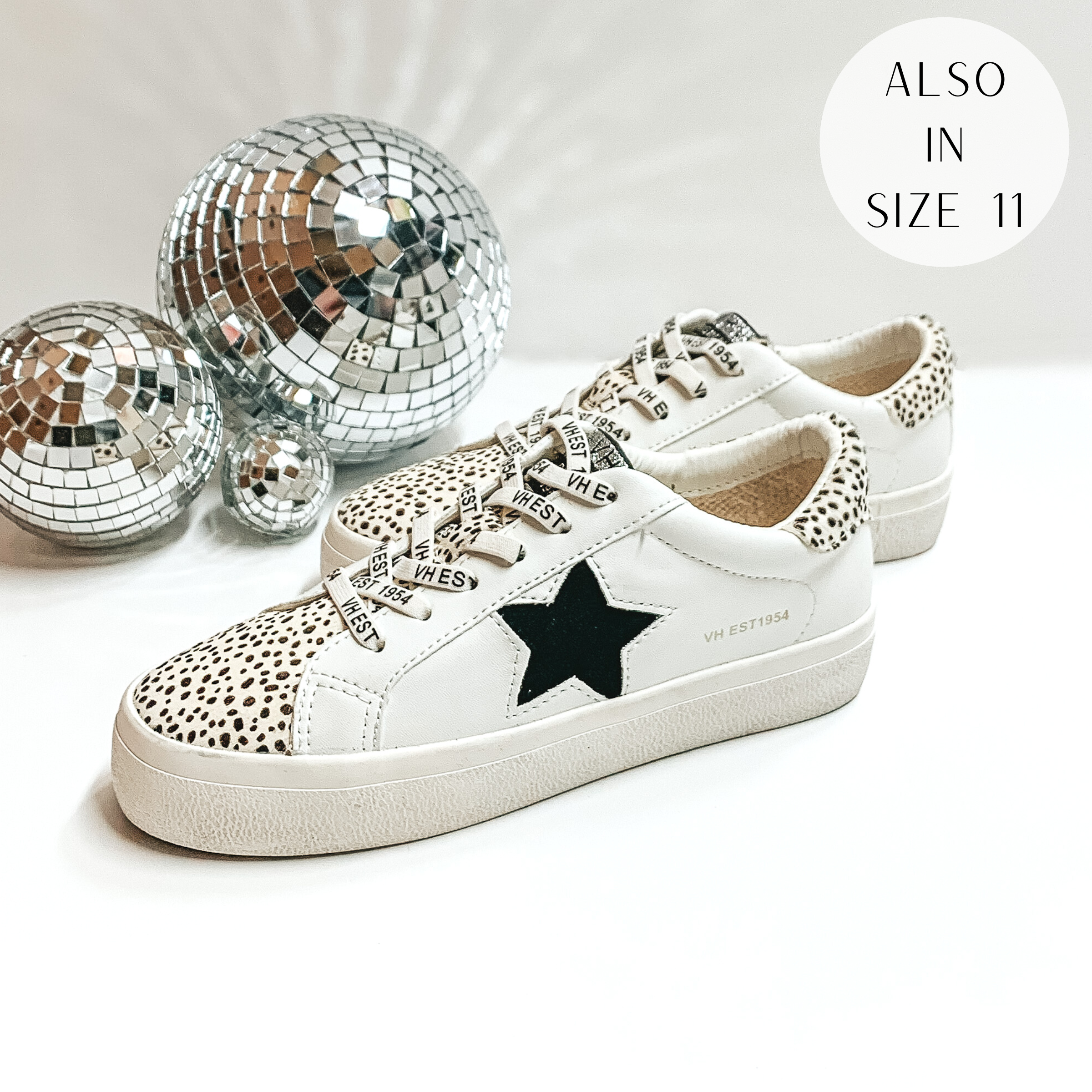 White tennis shoes with the tongue down to the toe part with a white and black cheetah print design. These shoes also have a black star emblem on the side of the shoe and a patch of white and black cheetah print on the heel. These shoes are pictured on a white background with disco balls on the left hand side.