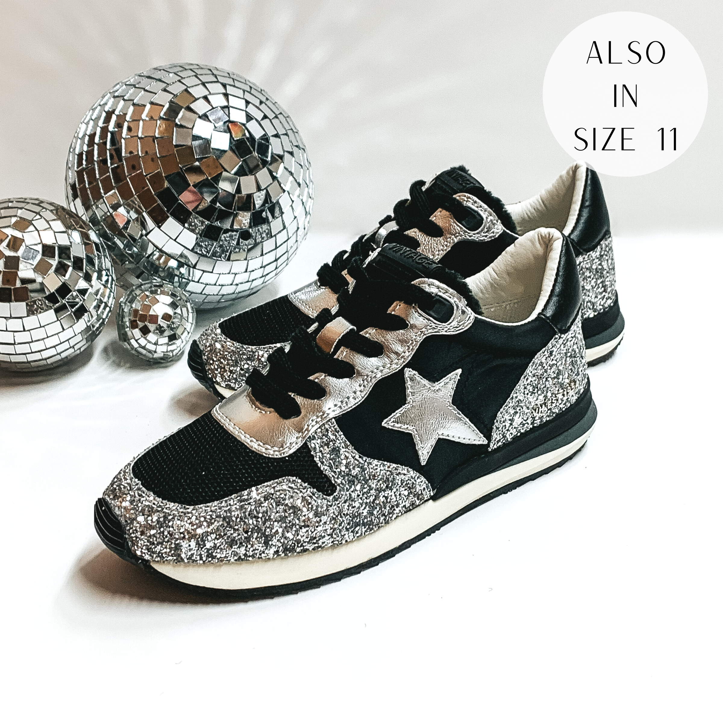 Black tennis shoes with silver glitter patches throughout. These shes also have a silver star emblem and a silver part around the black laces. These shoes are pictured on a white background with disco balls on the left hand side.