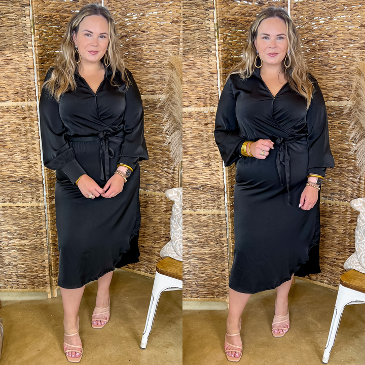 Model is wearing a black satin midi dress with long sleeves, a v neckline with a collar, and a waist tie. Model has it paired with nude heels and gold jewelry.