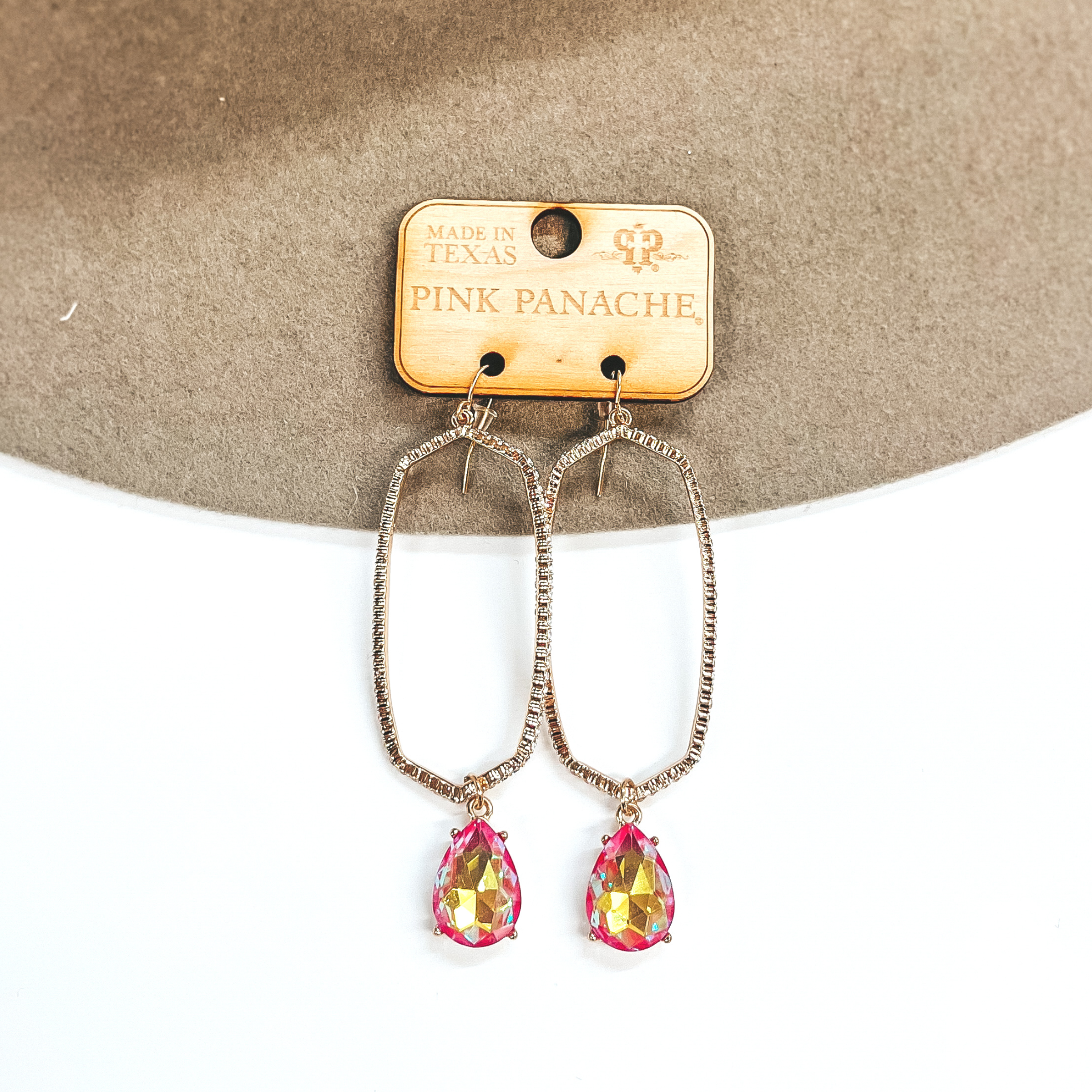 A pair of oval outline earrings that have crystals all the way around. These earrings have a coral AB crystal teardrop pendant at the bottom of each earring. Pictured on white background with a beige hat.